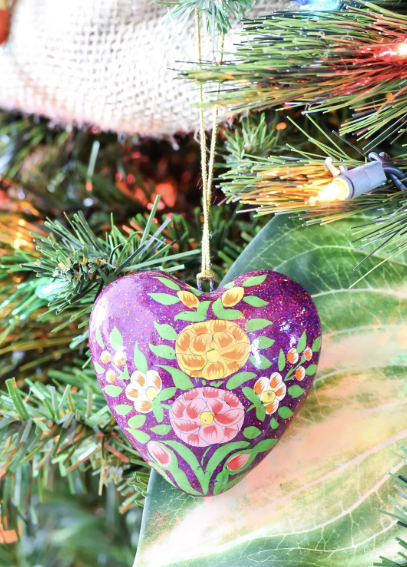 A heart-shaped ornament with colorful floral designs by Crimson Design Group hangs on a Christmas tree, surrounded by green branches and holiday lights.