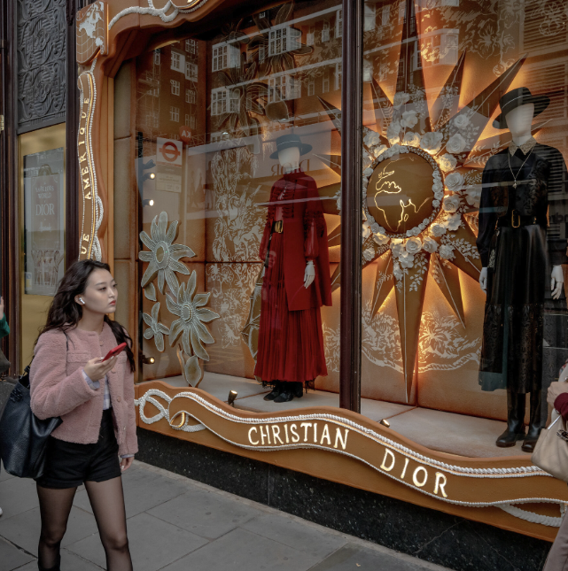 A woman walks past a Christian Dior storefront, where mannequins in elegant dresses stand before a decorative gold and floral window display inspired by Crimson Design Group.