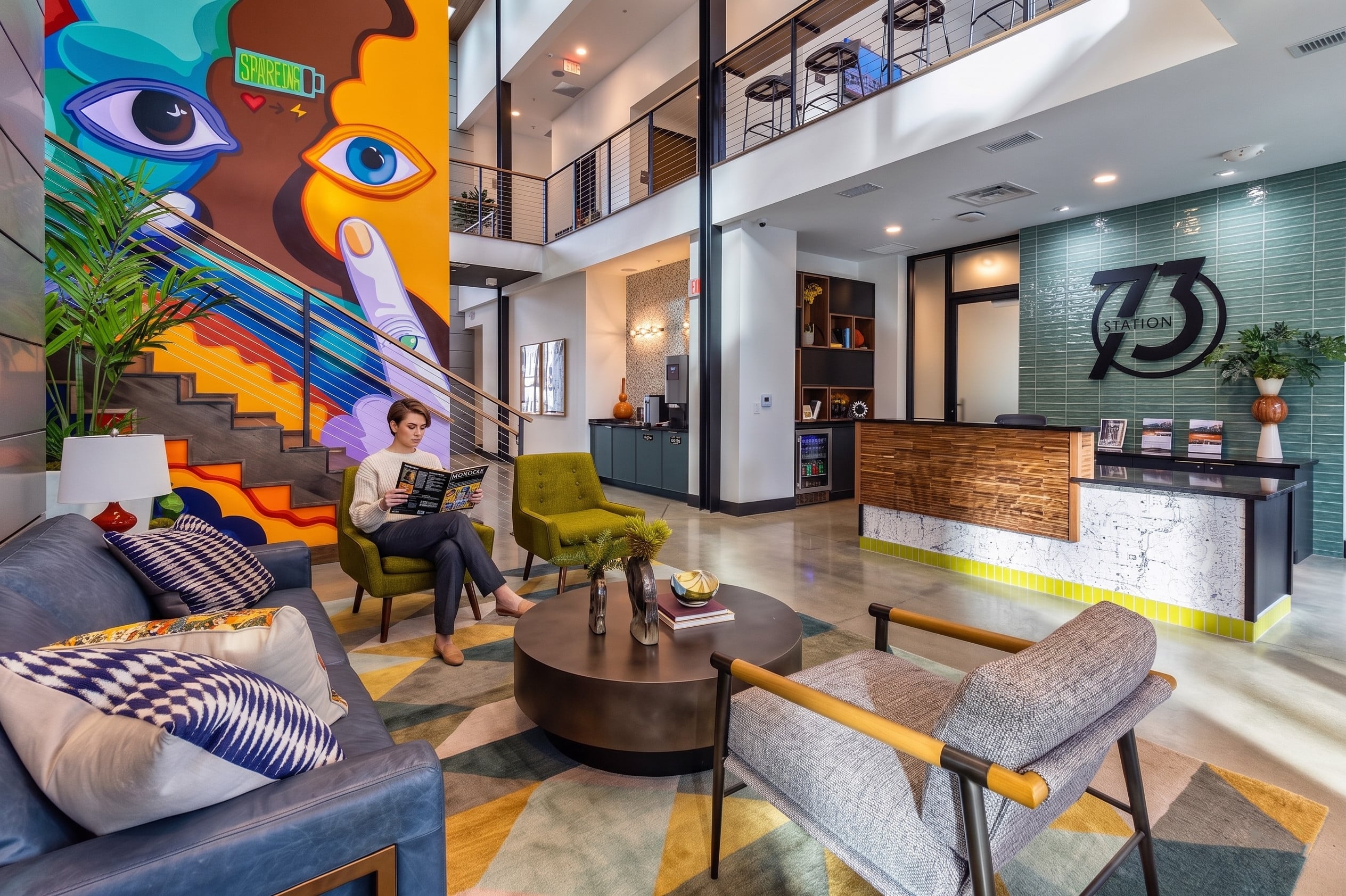 Modern lobby with colorful wall mural, contemporary furniture, and a reception desk. A woman sits reading a magazine on a sofa.