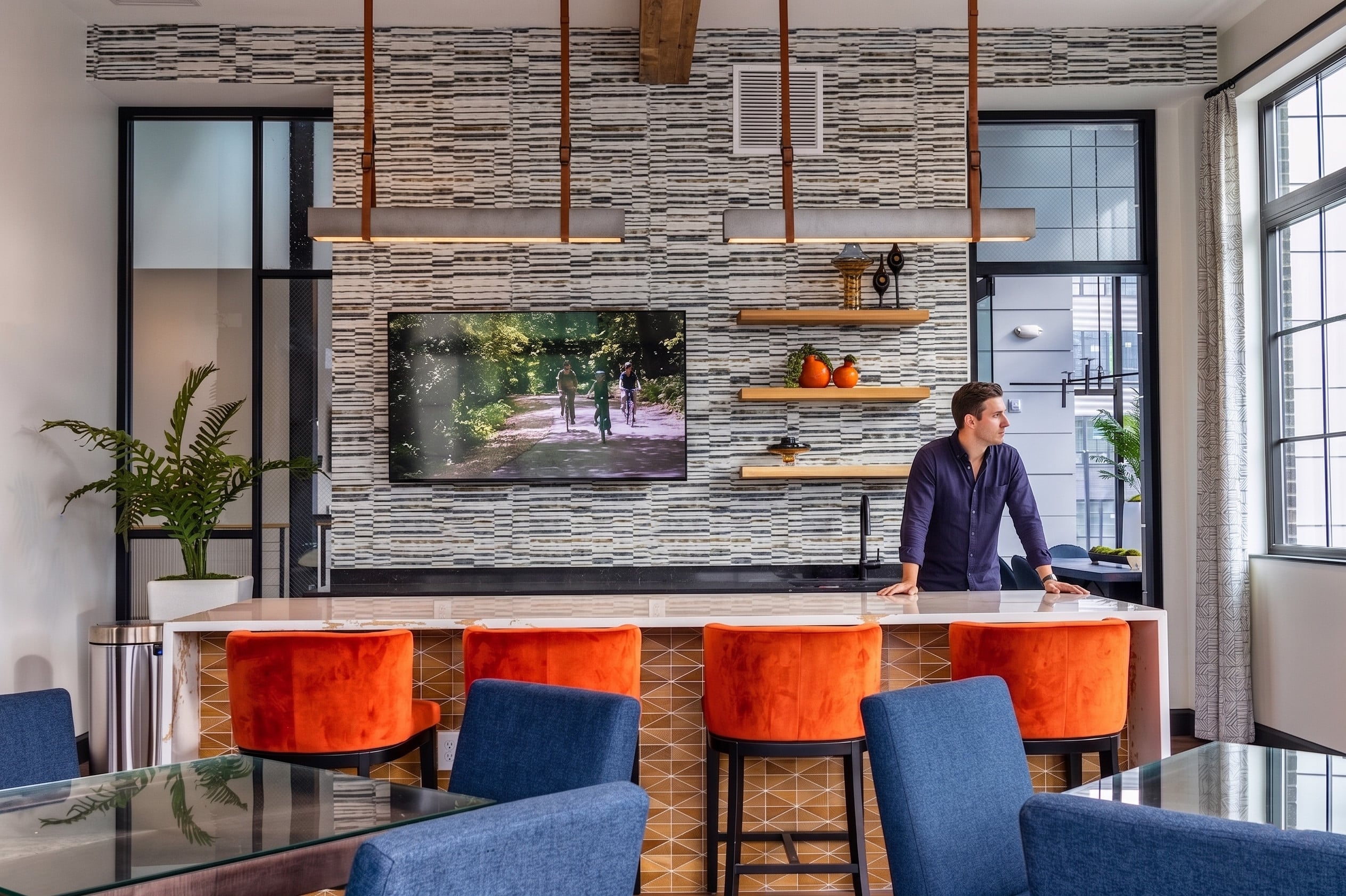 A man stands at a kitchen island with orange barstools in a modern, well-lit room featuring a TV, open shelves with decor, and large windows.