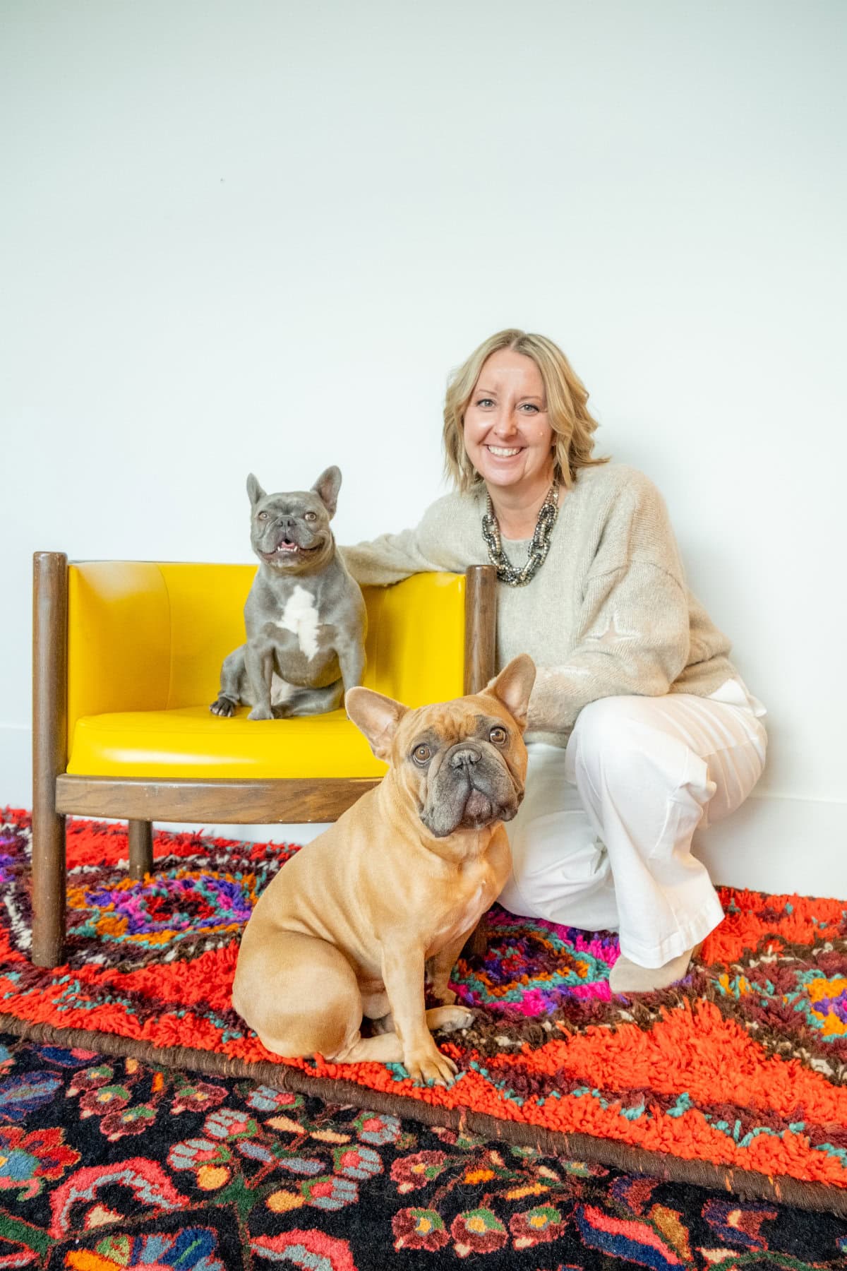 A woman kneels beside a yellow chair with a gray French bulldog sitting on it and a tan French bulldog sitting on a colorful rug in front.