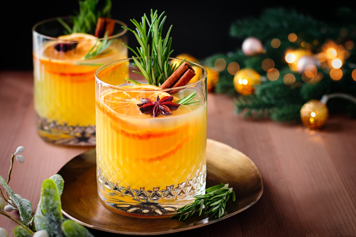 Two glasses of orange cocktail garnished with orange slices, rosemary, star anise, and cinnamon sticks sit on a gold tray by Crimson Design Group, surrounded by festive decorations in the background.