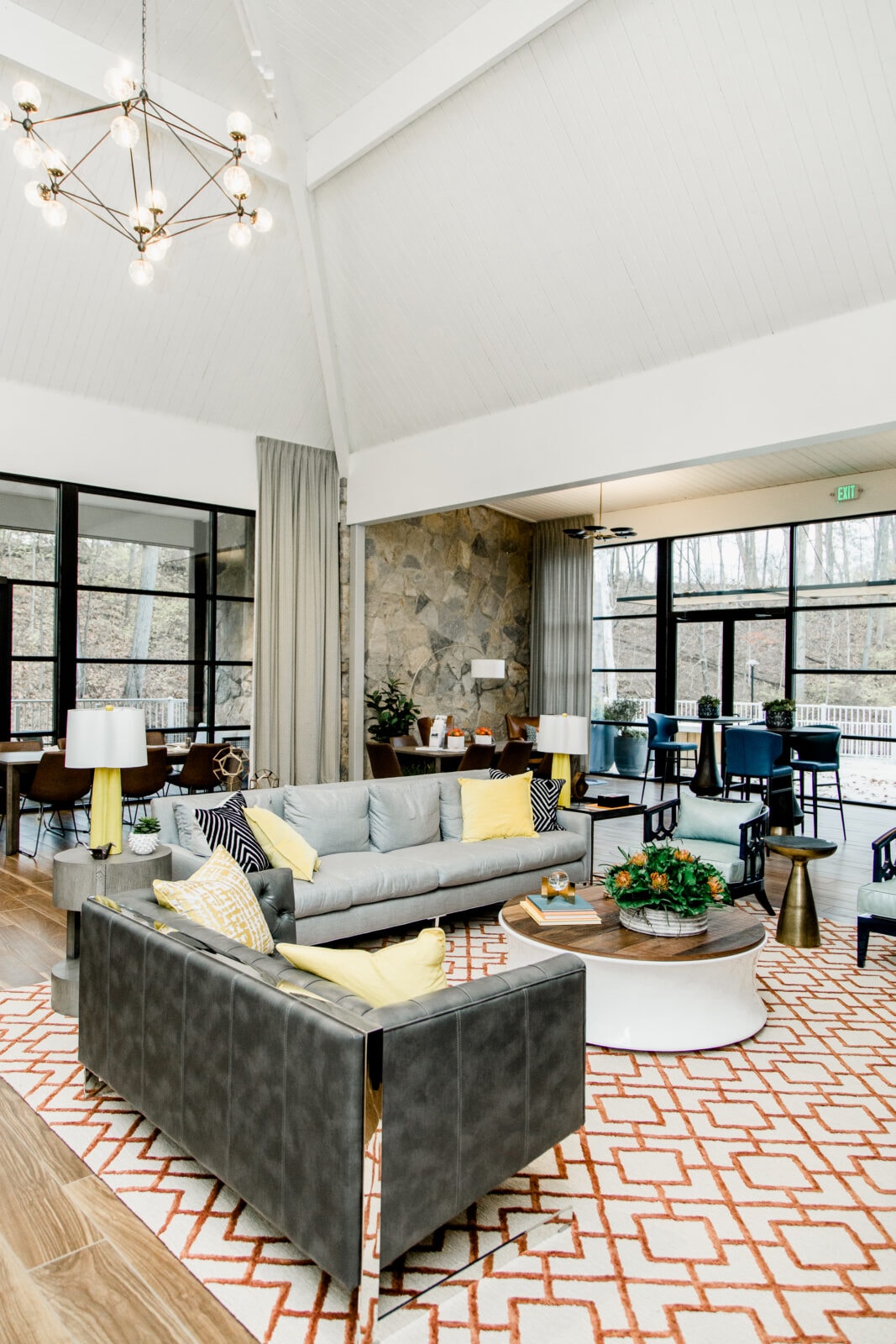 A modern living room with gray sofas, yellow and black cushions, a geometric rug, a round coffee table, large windows, and a stone accent wall under a vaulted white ceiling.