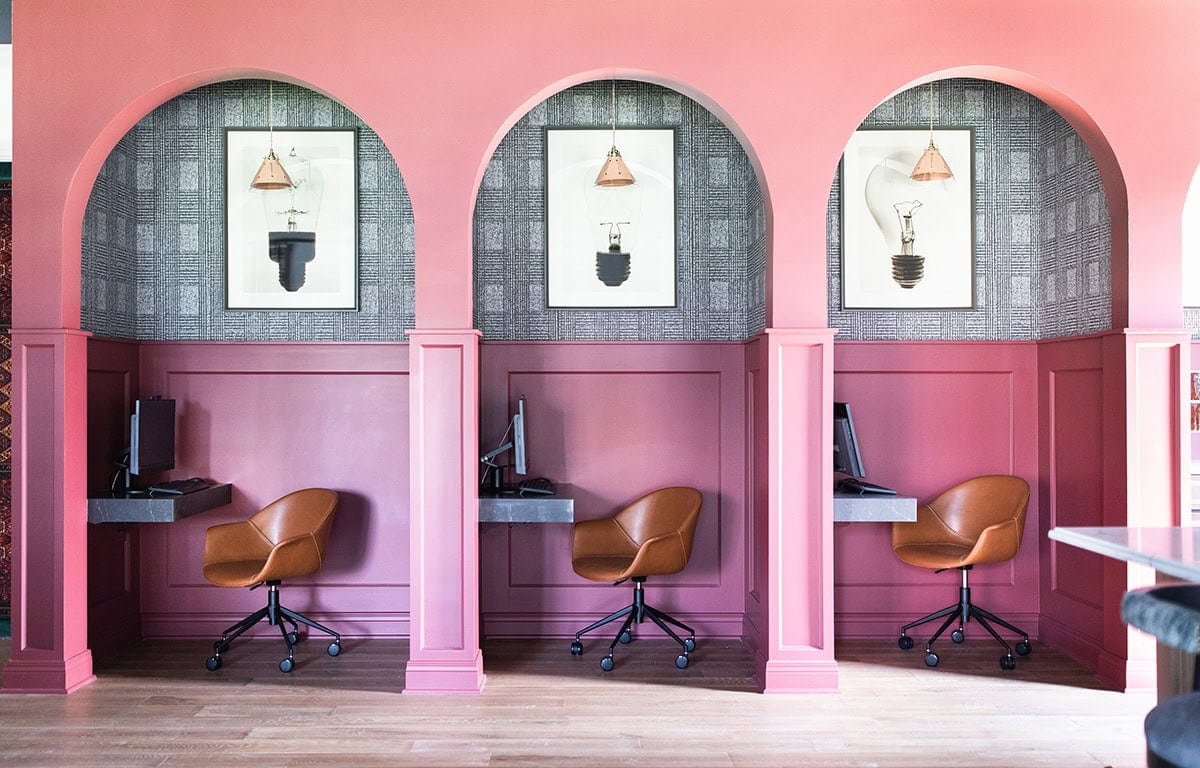 Three workstations with brown chairs and desktop computers are set in pink alcoves, each with a framed light bulb print above and geometric wallpaper backgrounds.
