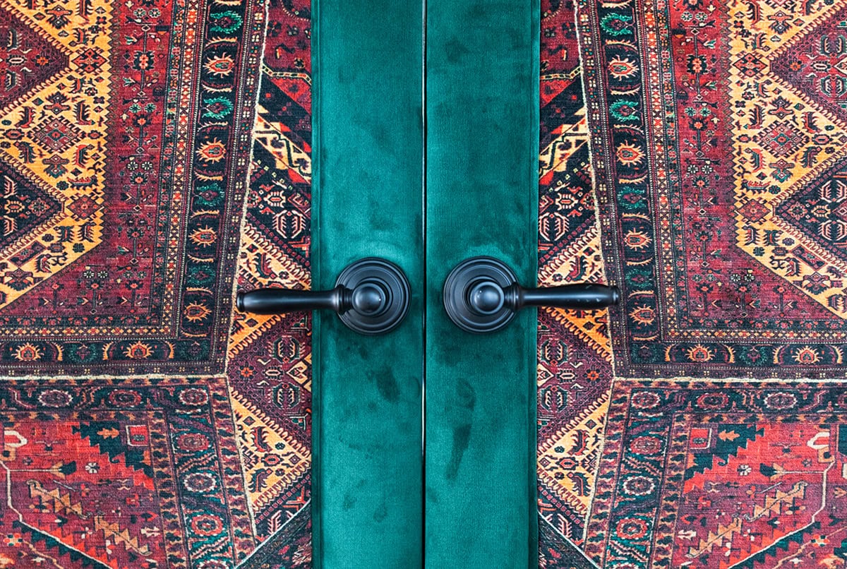 Two green doors with black handles against a vibrant, patterned, red and yellow rug or tapestry backdrop.