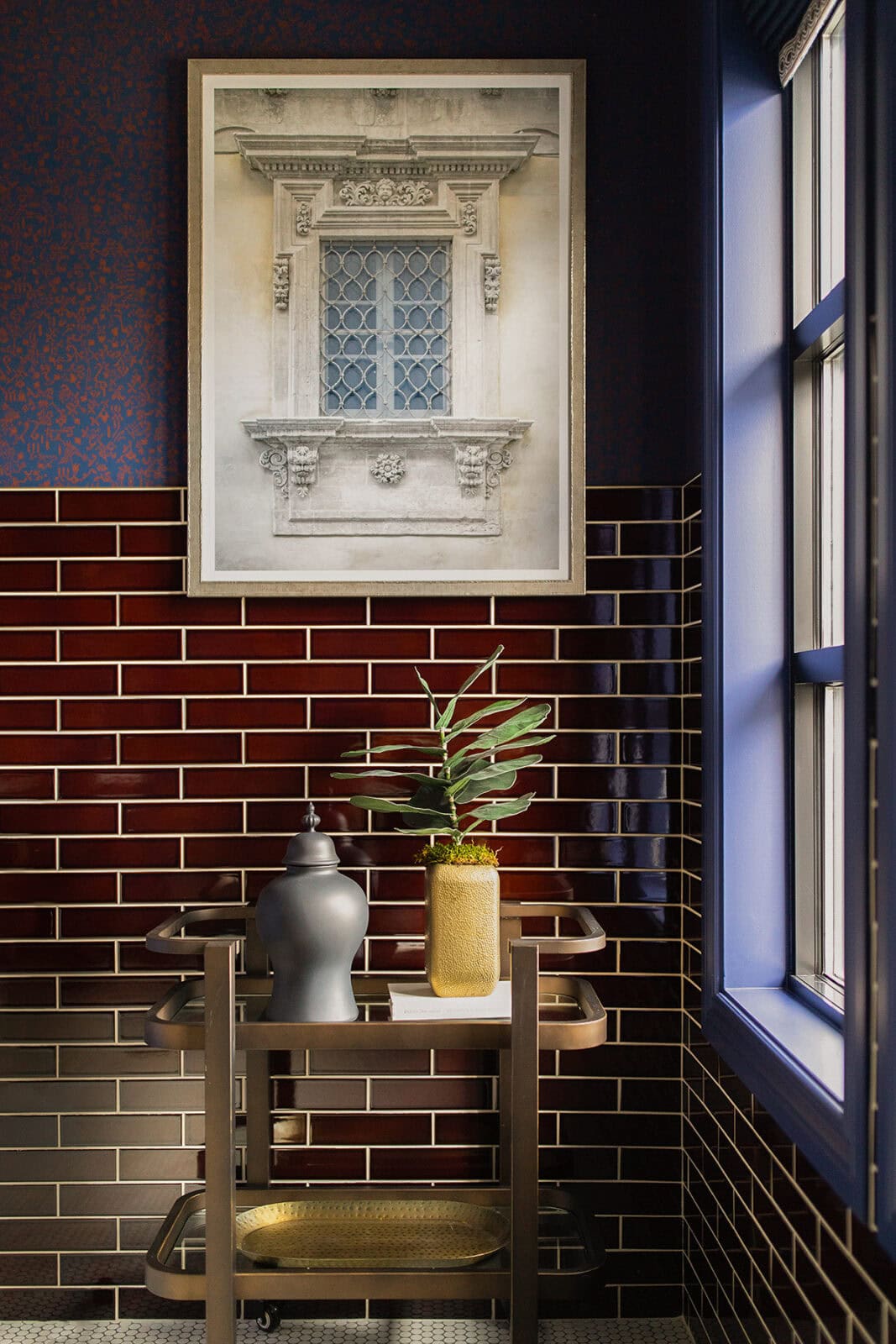 A framed architectural print hangs above a bar cart with a black vase, a yellow vase holding a leafy plant, set against dark brown subway tiles and a window with blue trim.