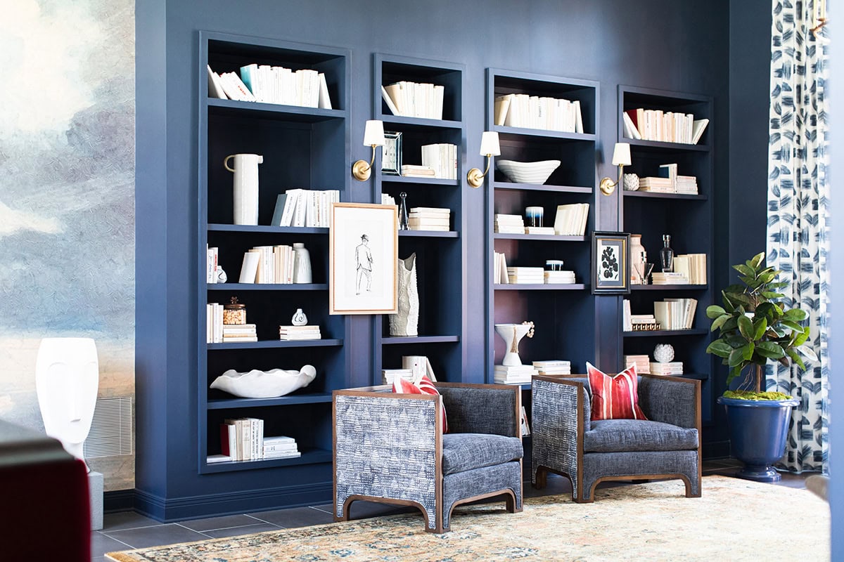 Three navy blue bookshelves filled with white books and decorative items stand behind two gray armchairs with red pillows; a potted plant sits nearby on a patterned rug.