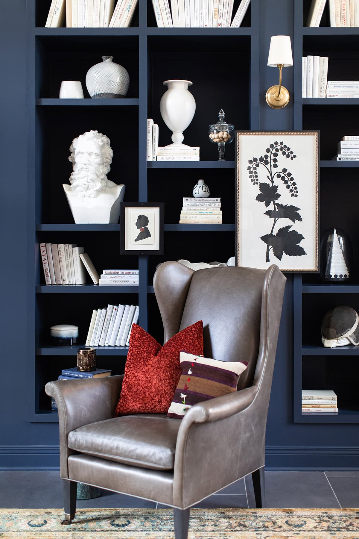 A gray leather armchair with red and striped pillows sits in front of a dark blue built-in bookshelf filled with books, vases, art, and decor items.