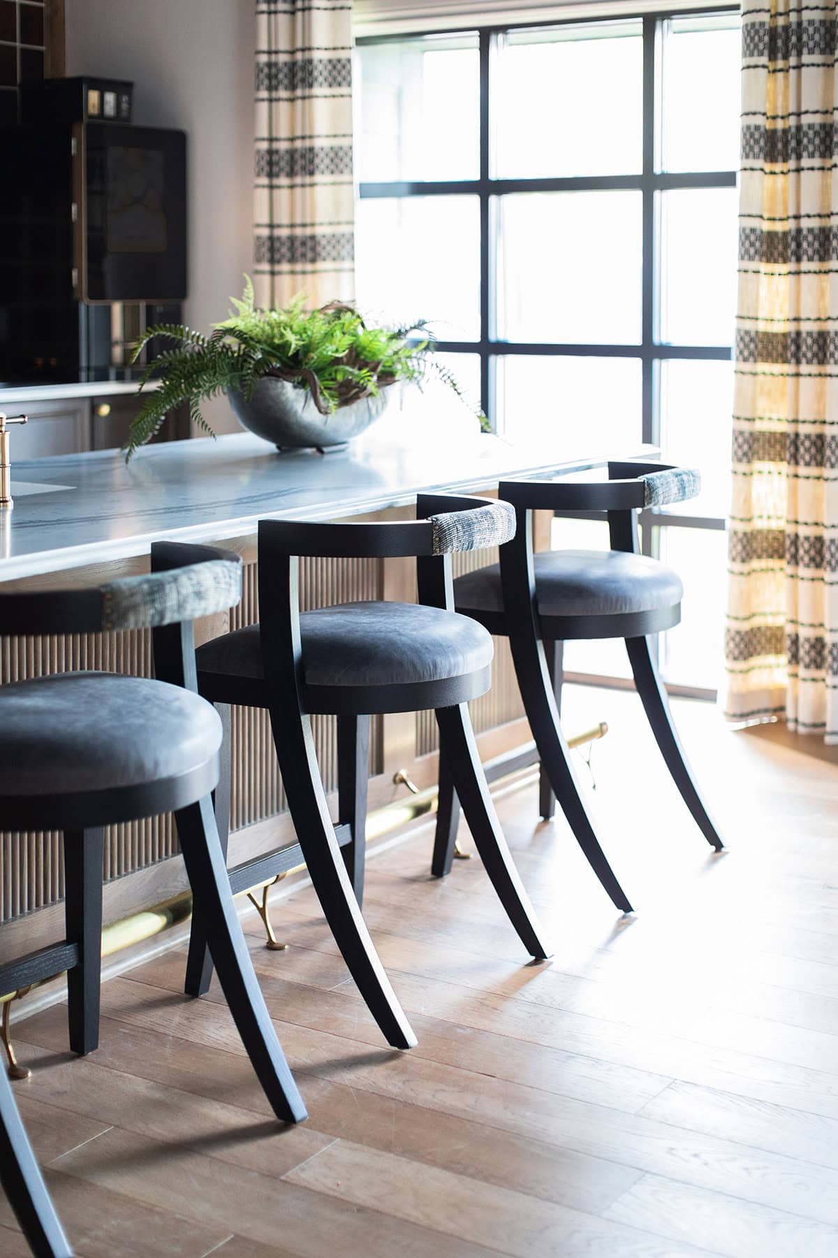 A modern kitchen with a marble countertop, three black barstools, a bowl of ferns, large windows, and patterned yellow curtains.