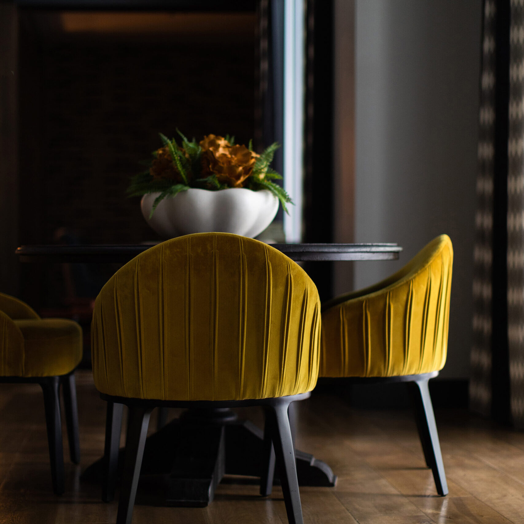 A dark dining area by Crimson Design Group features a round table with a white bowl of plants as the centerpiece and yellow velvet chairs on a wooden floor.