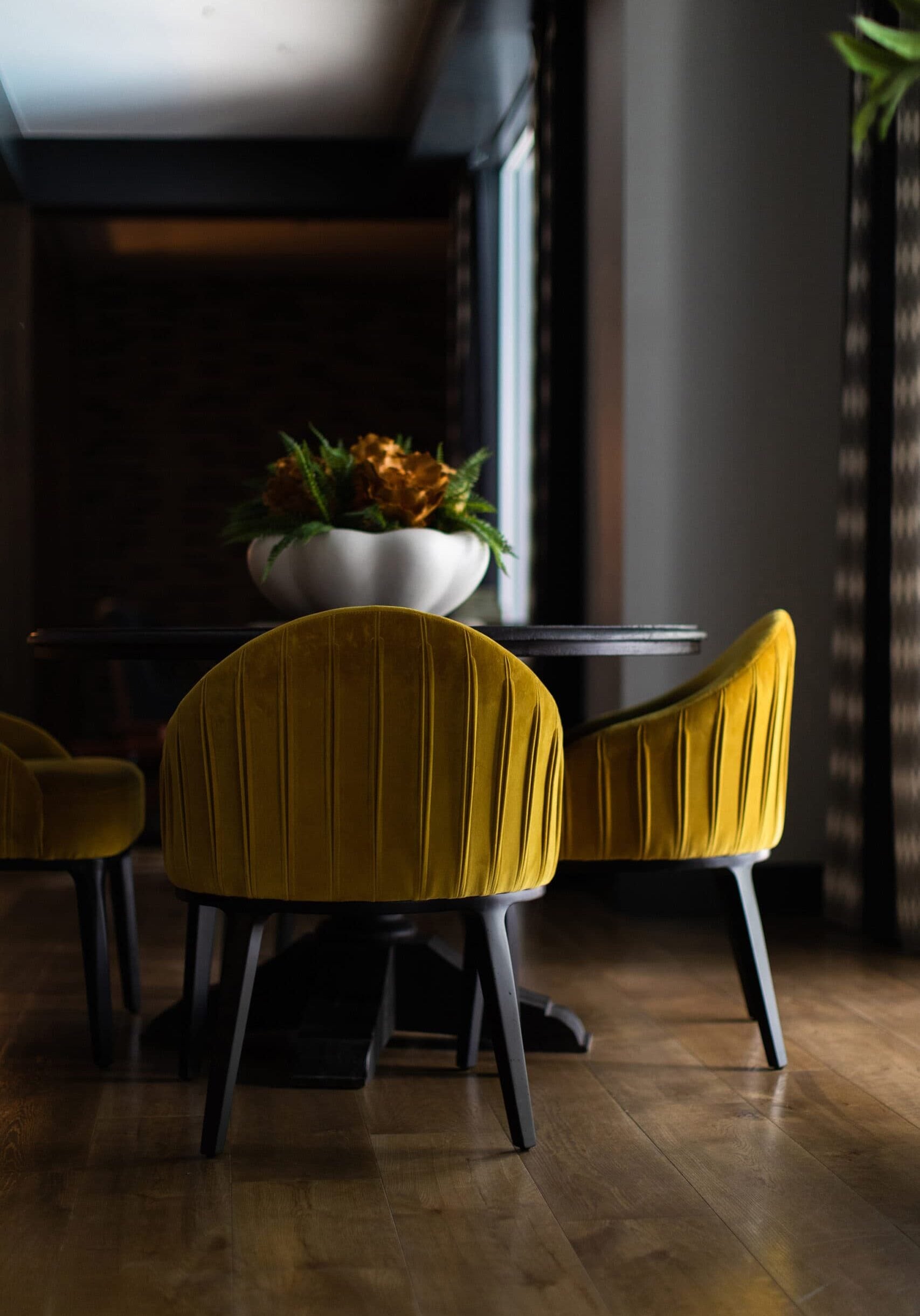 A dining table with a large white bowl of plants, surrounded by mustard yellow velvet chairs on a wooden floor in a dimly lit room.