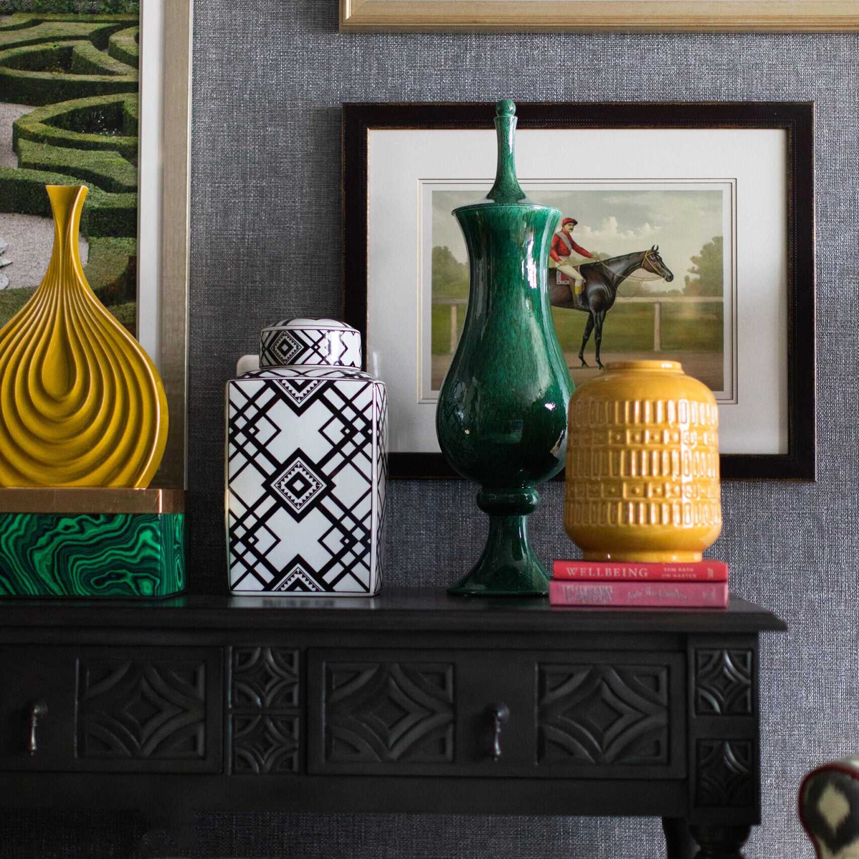 A black table by Crimson Design Group displays decorative vases and books, with framed artwork and photographs hanging on a textured gray wall behind it.