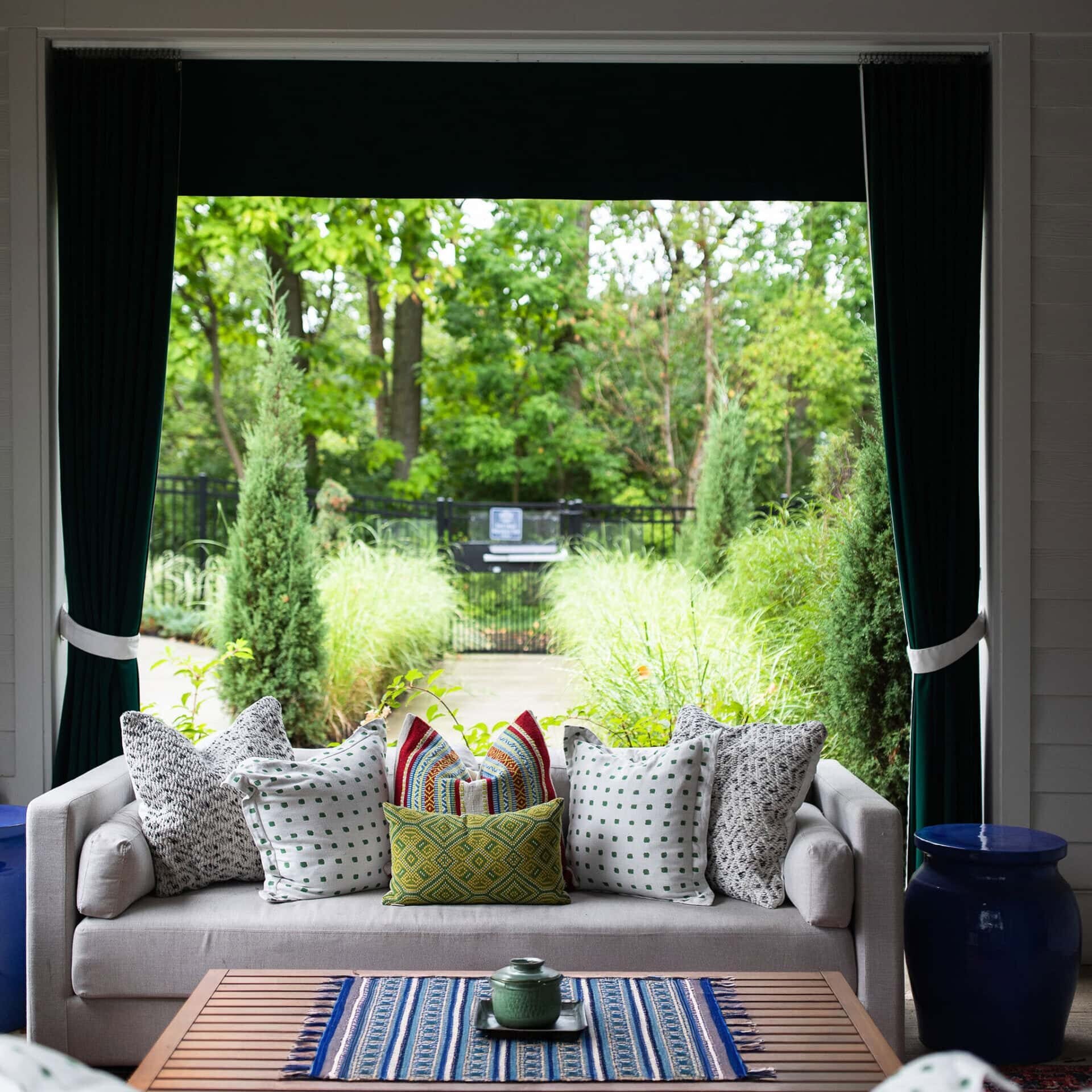 A covered patio with a beige sofa, patterned pillows, a wooden coffee table, and green plants visible through an open window.