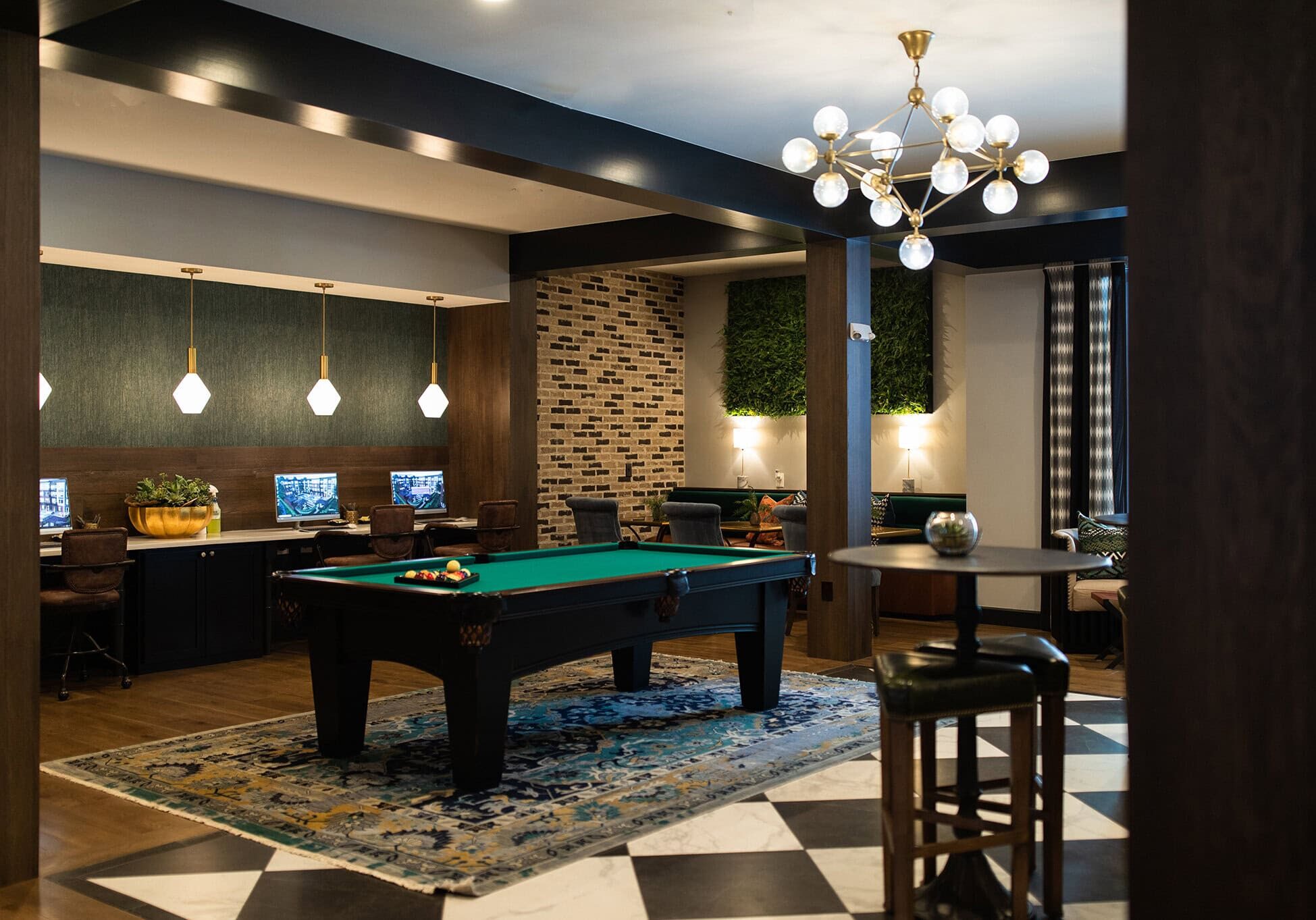 A modern lounge with a pool table on a rug, high-top table with stools, computer workstations, pendant lights, and contemporary decor.