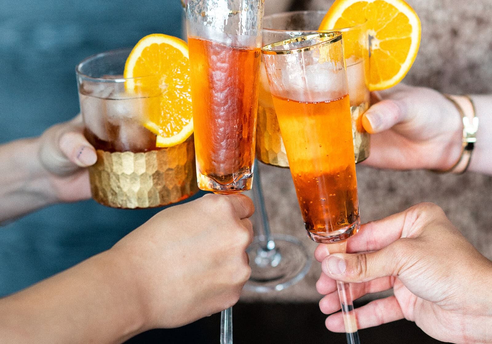 Four people clinking glasses filled with iced cocktails garnished with orange slices.