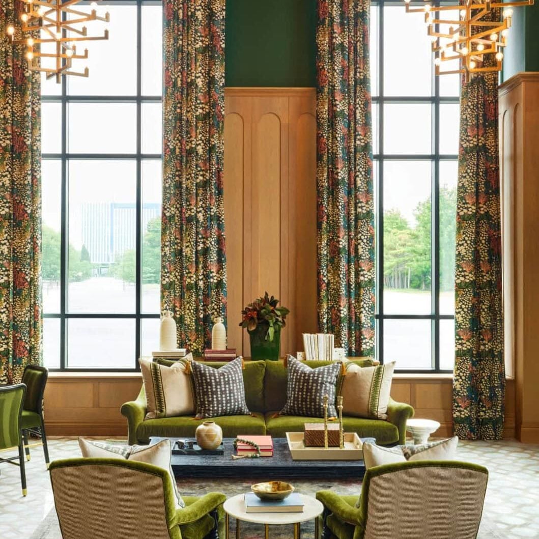 A high-ceiling multi-family amenity lobby featuring tall arched windows, deep green walls, velvet olive-green furniture, and geometric gold chandeliers.