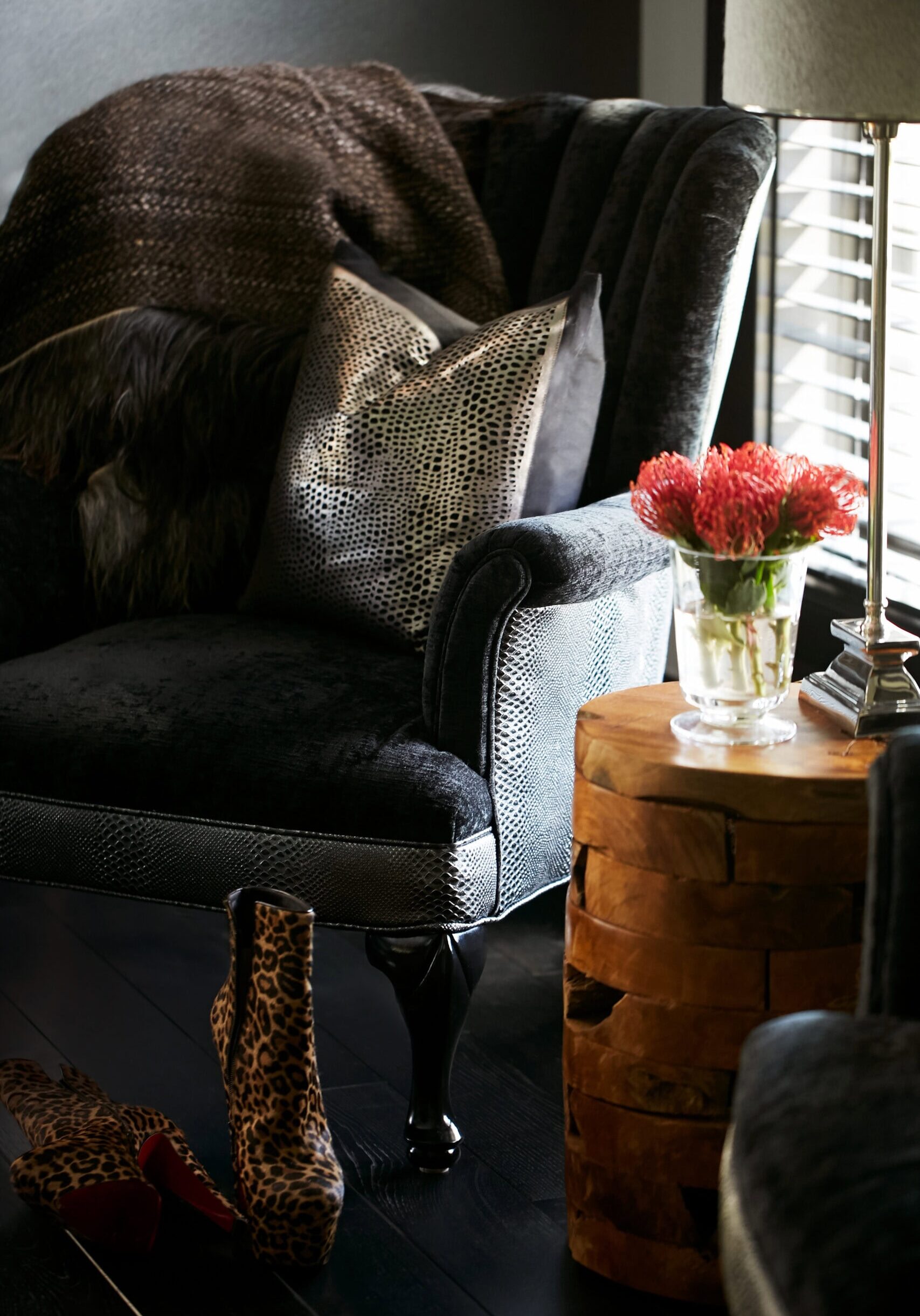 A black armchair with a metallic pillow and throw sits next to a wooden side table with a glass vase of pink flowers; leopard print shoes are on the dark floor.