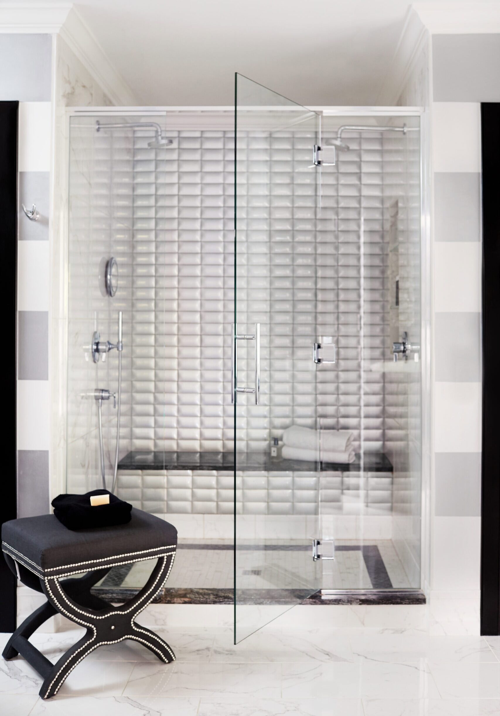 Modern glass shower with white textured tile walls, dual showerheads, a built-in bench with folded towels, and a black ottoman with silver studs outside.
