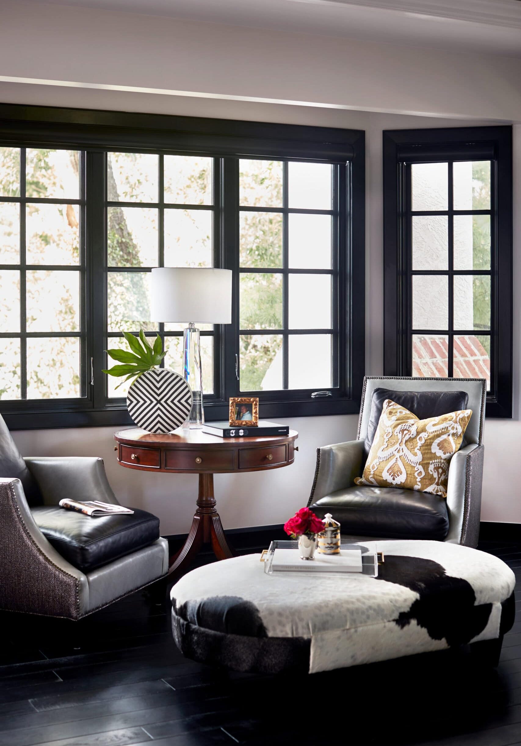 A modern sitting area with two black armchairs, a round ottoman, a wooden side table with a lamp and vase, and large windows framed in black.