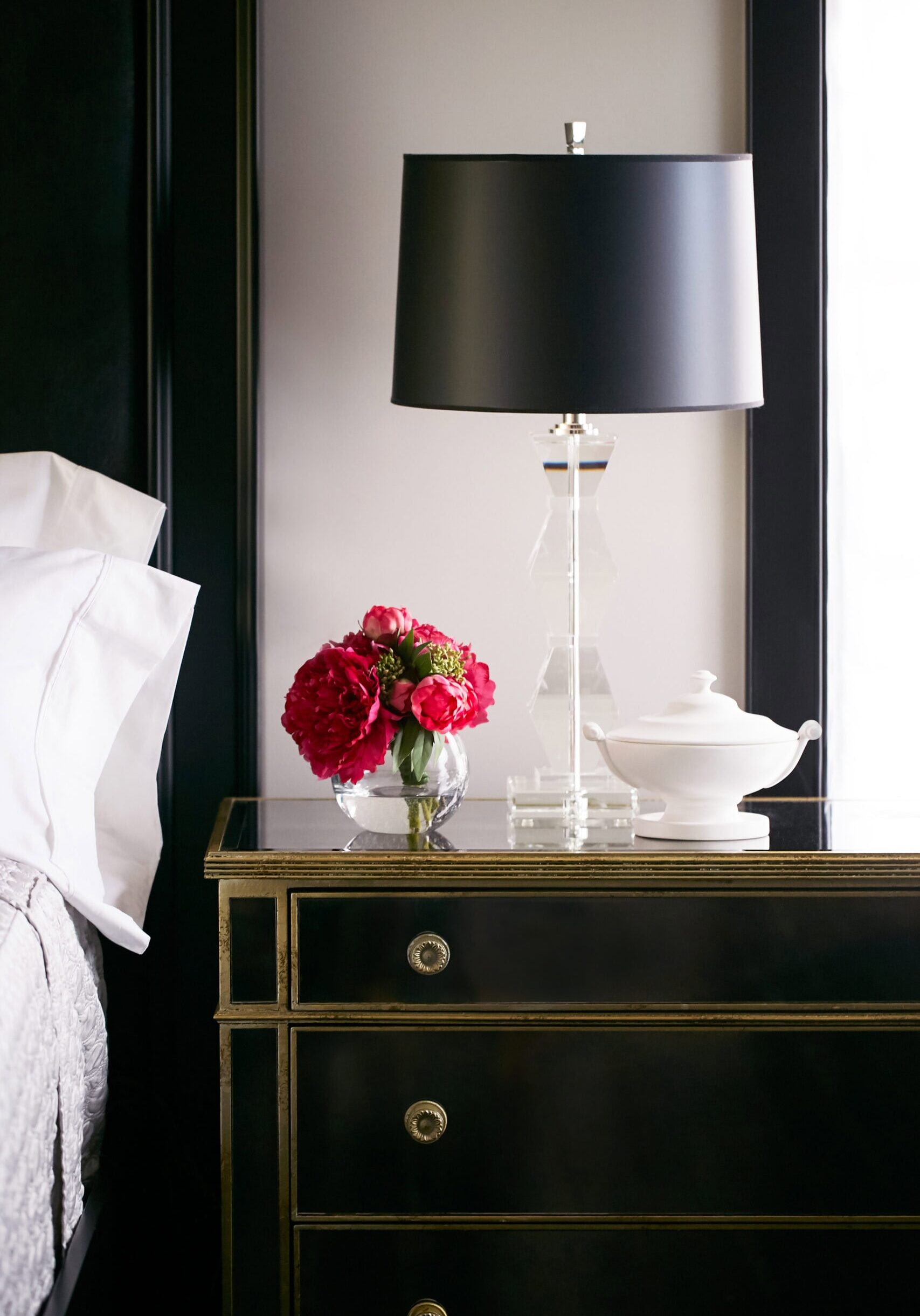A glass lamp, a vase with red flowers, and a white dish sit on a black nightstand beside a bed with white pillows.
