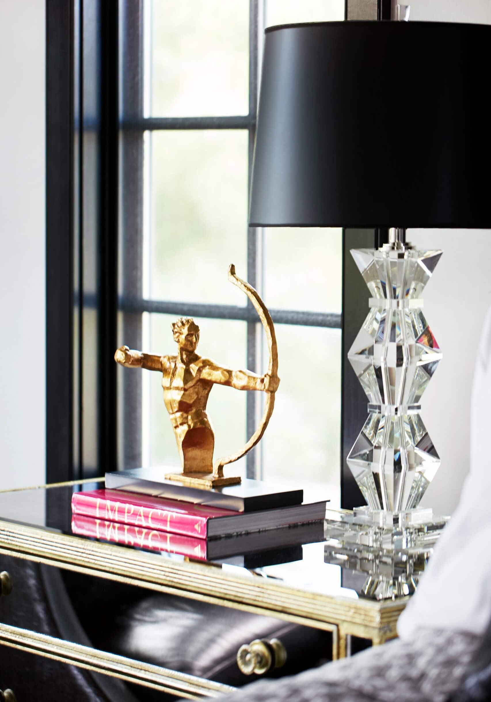A gold archer figurine sits atop stacked books on a mirrored nightstand beside a geometric crystal lamp with a black shade, near a window.