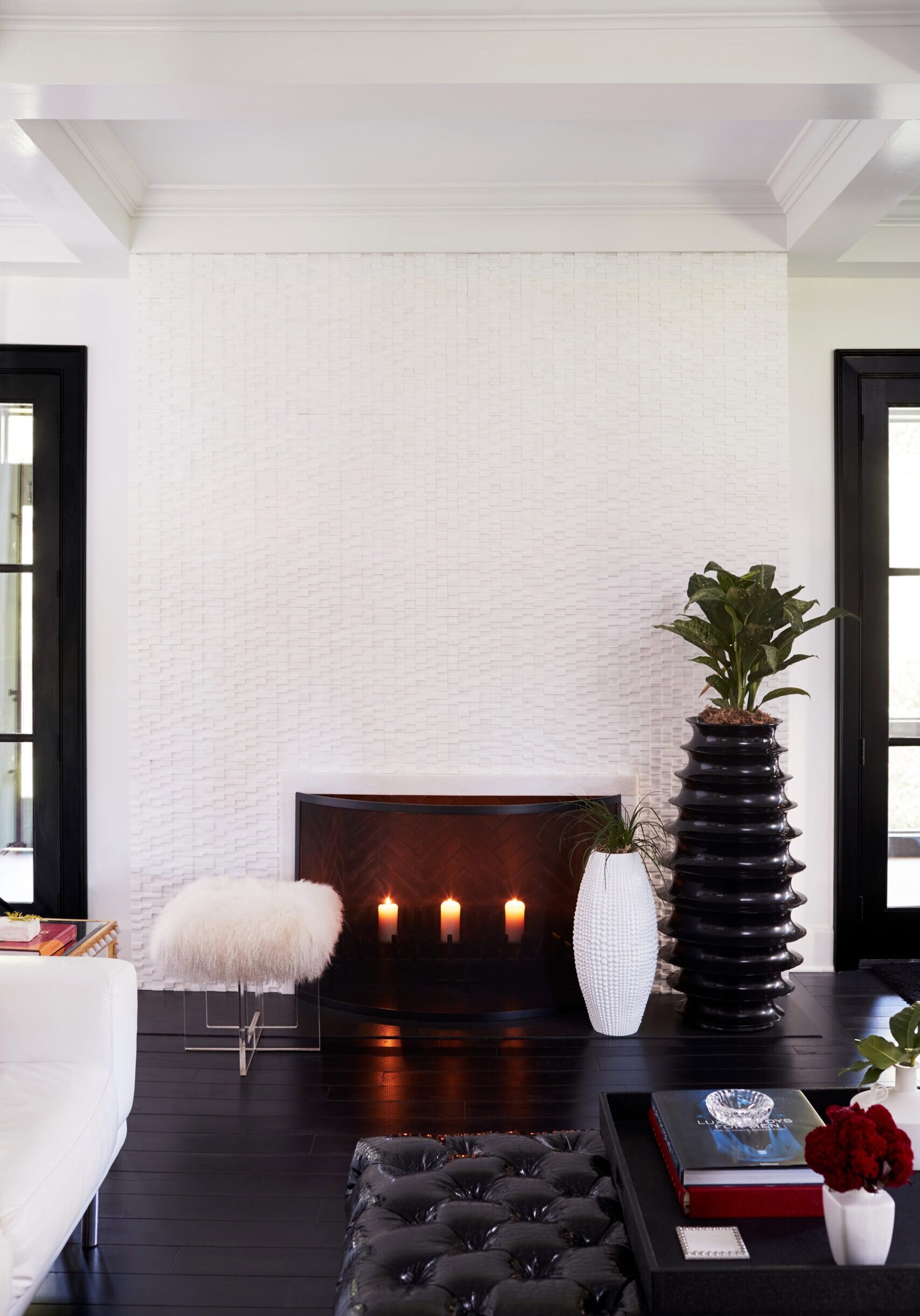 A modern living room with a white furry stool, black ottoman, candles in a fireplace, tall black vase with a plant, and white walls with black-trimmed windows.