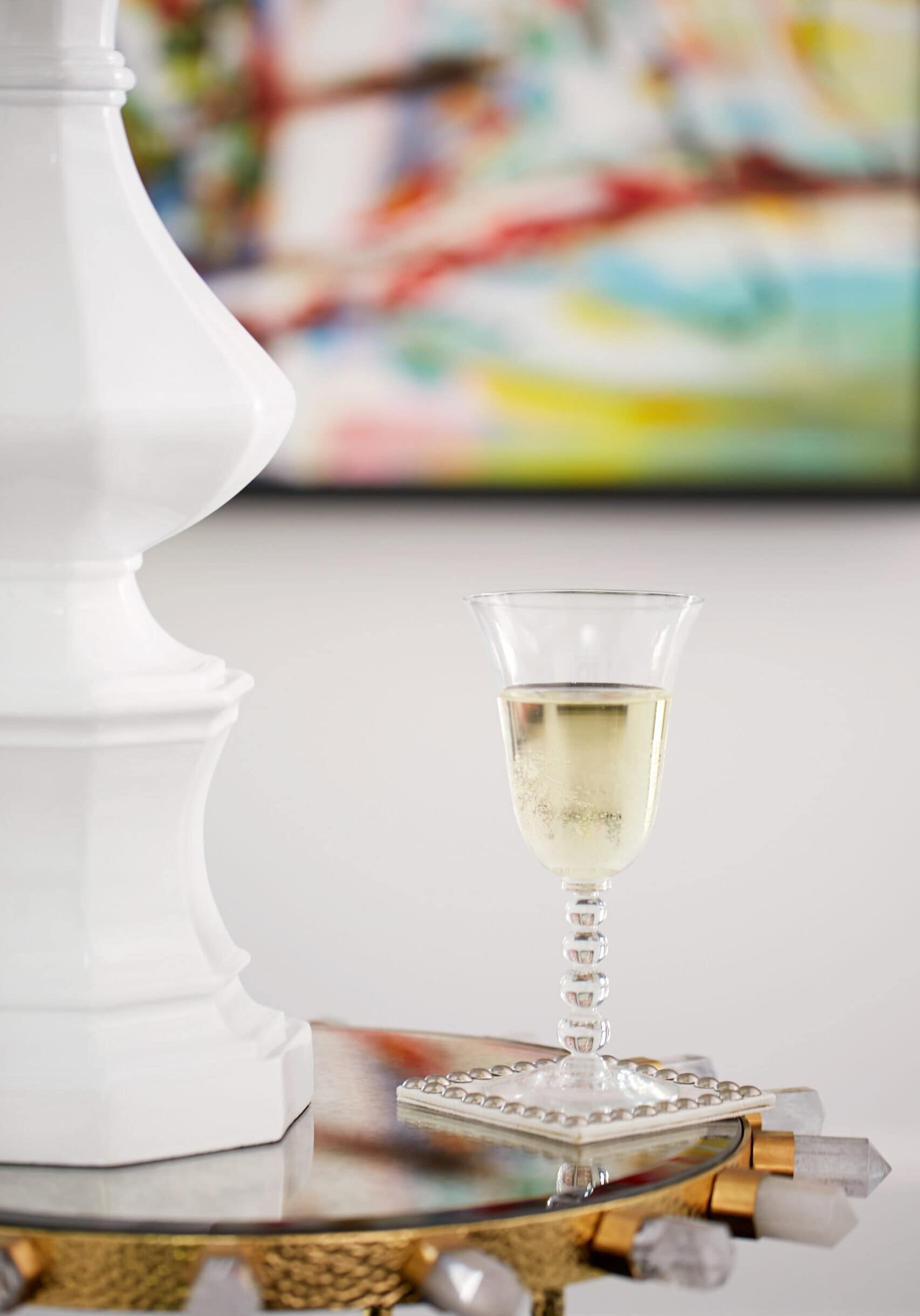 A glass of white wine sits on a coaster on a reflective table next to a large white decorative object, with a colorful abstract painting in the background.