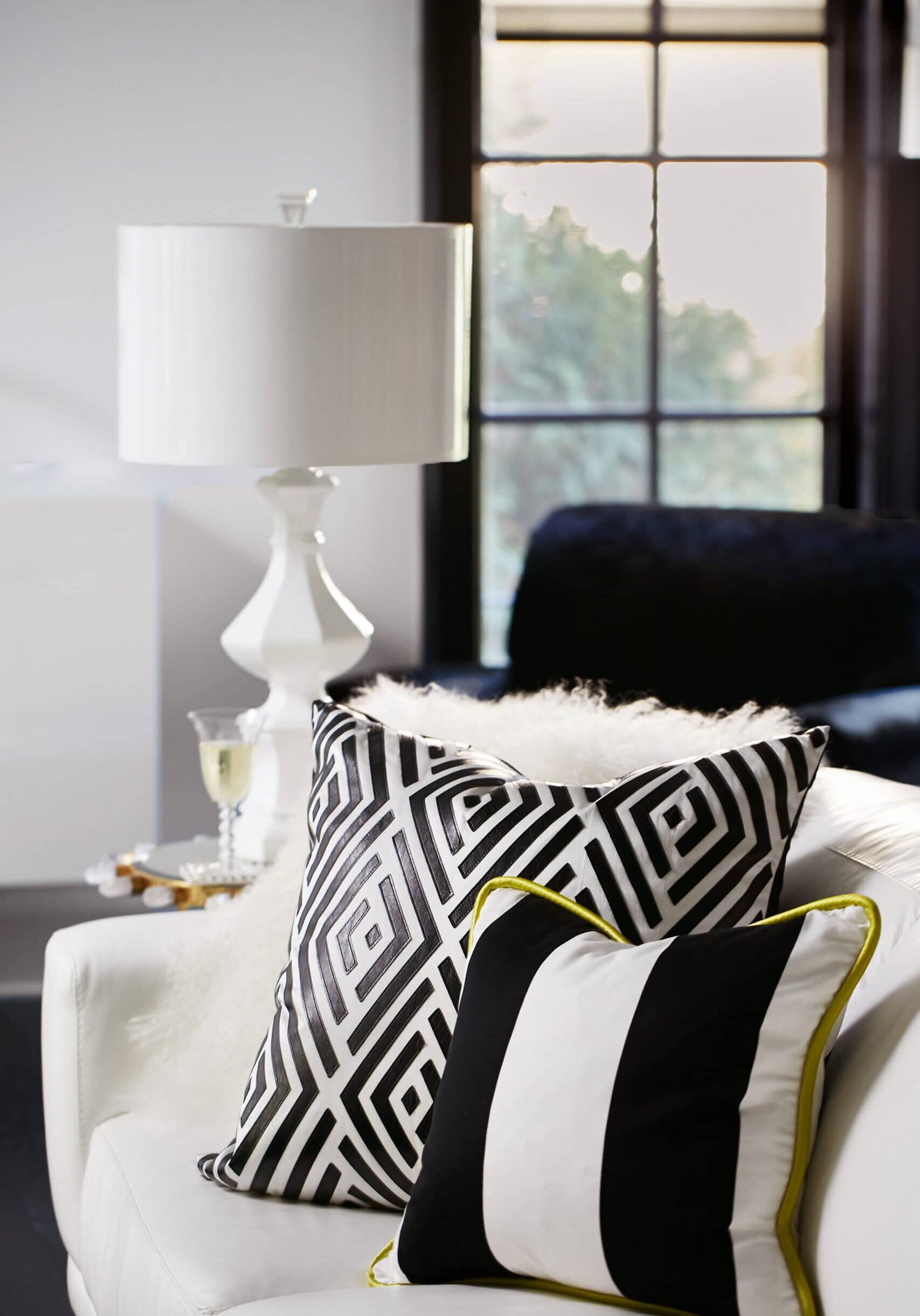 White sofa with two black-and-white patterned pillows, a white table lamp, and a window in the background. A glass of white wine sits on a side table.