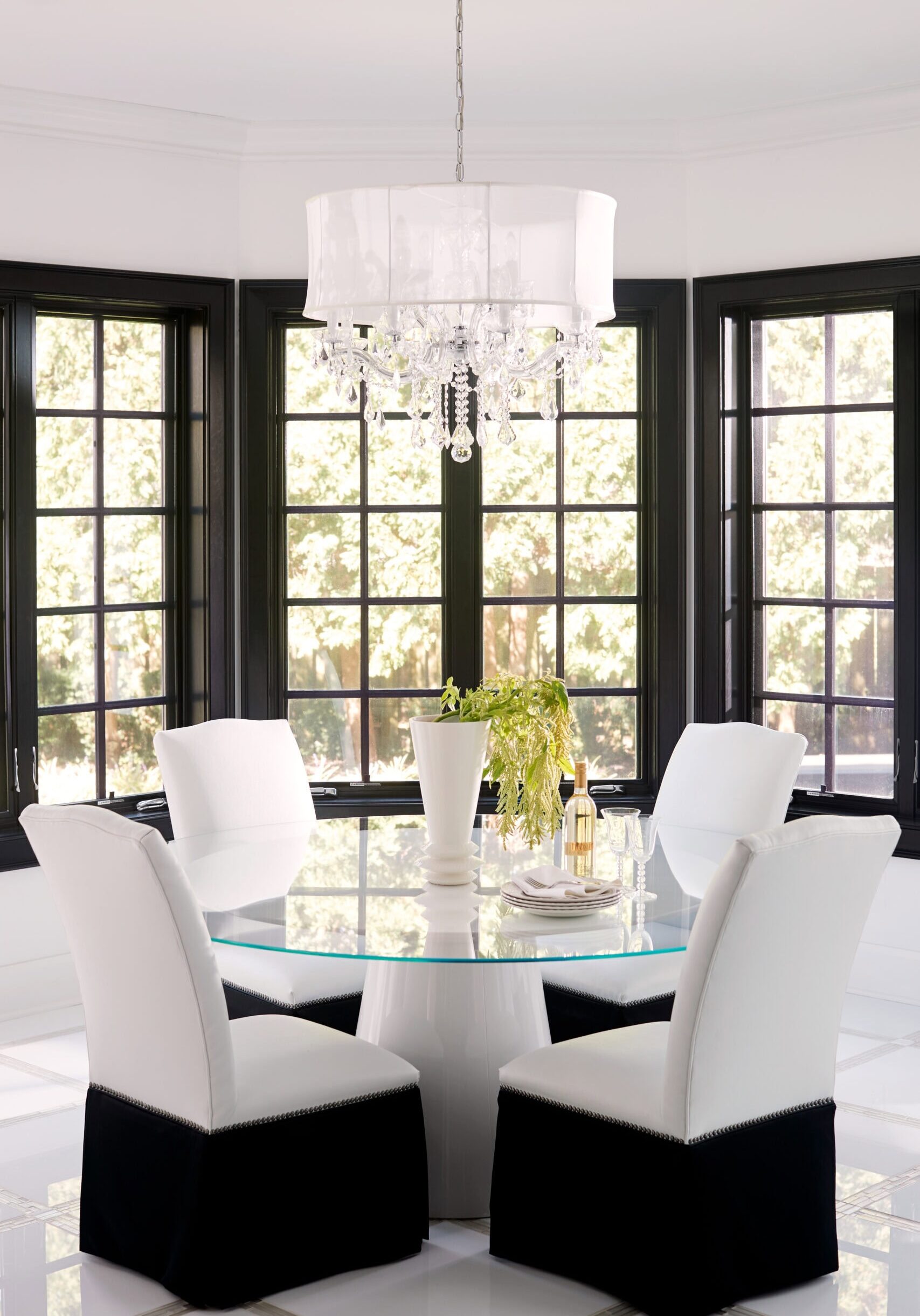 Modern dining room with a round glass table, four white chairs with black bases, a white vase with greenery, wine bottle, and a crystal chandelier, surrounded by black-framed windows.
