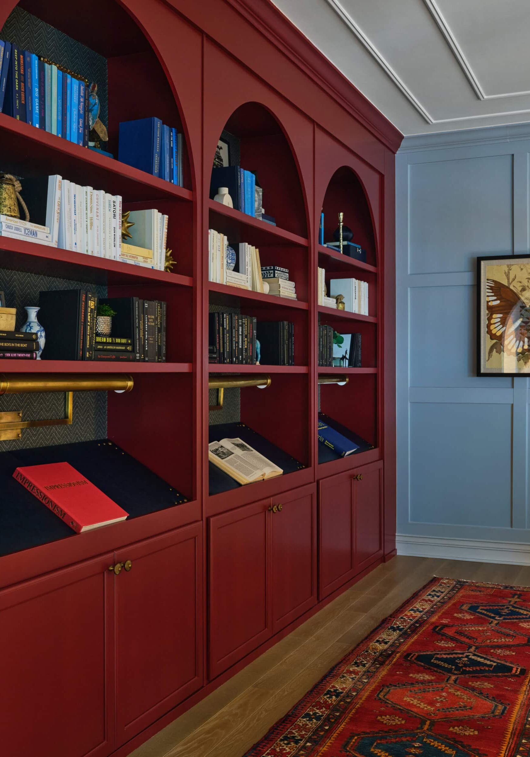 Floor-to-ceiling red bookshelves with books and decor stand against a wall in a room with blue paneling, wood flooring, and a patterned rug. A framed bird artwork hangs on the blue wall.