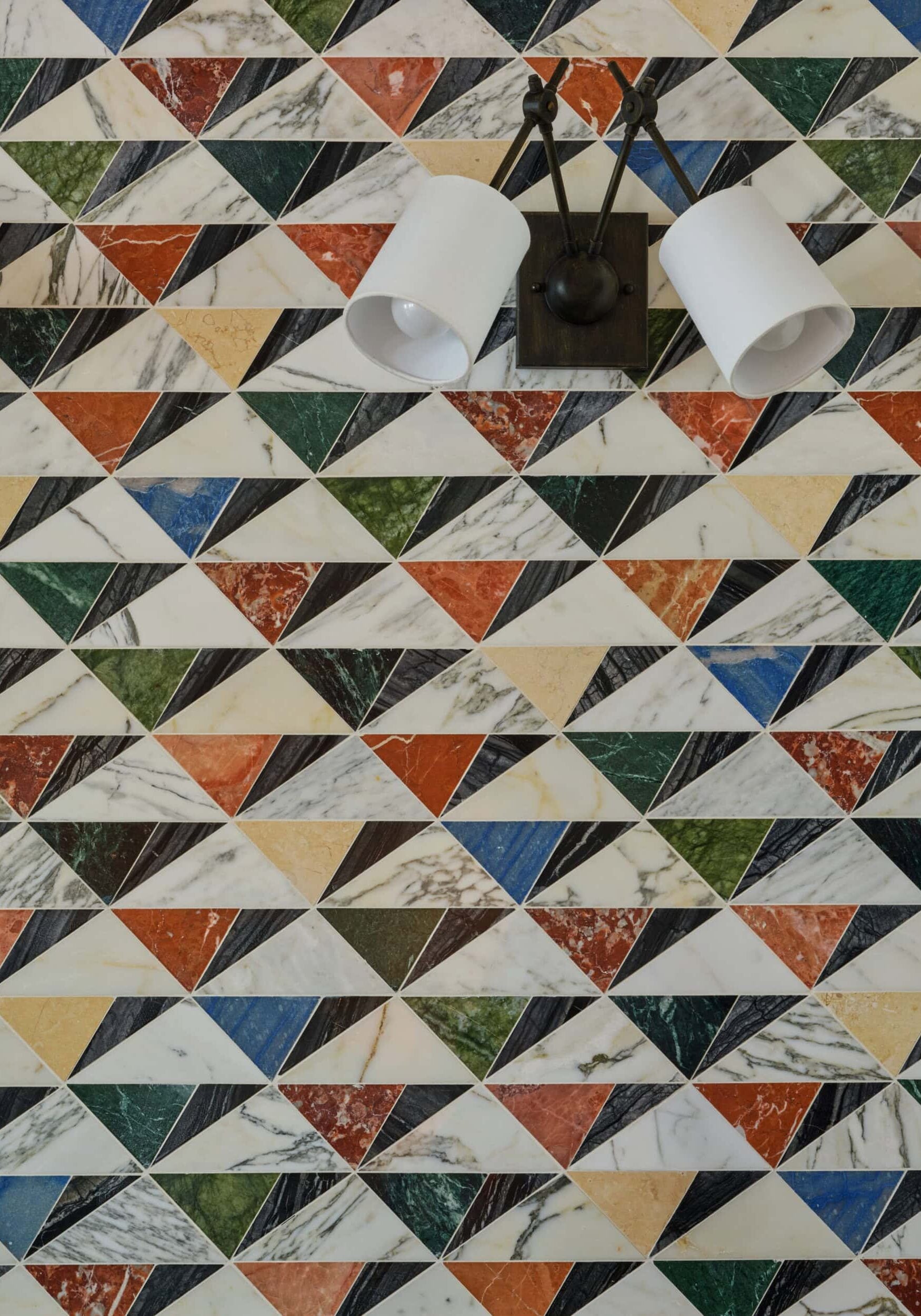 A wall with a geometric pattern of multicolored marble triangles, with a modern, double white sconce mounted near the top center.