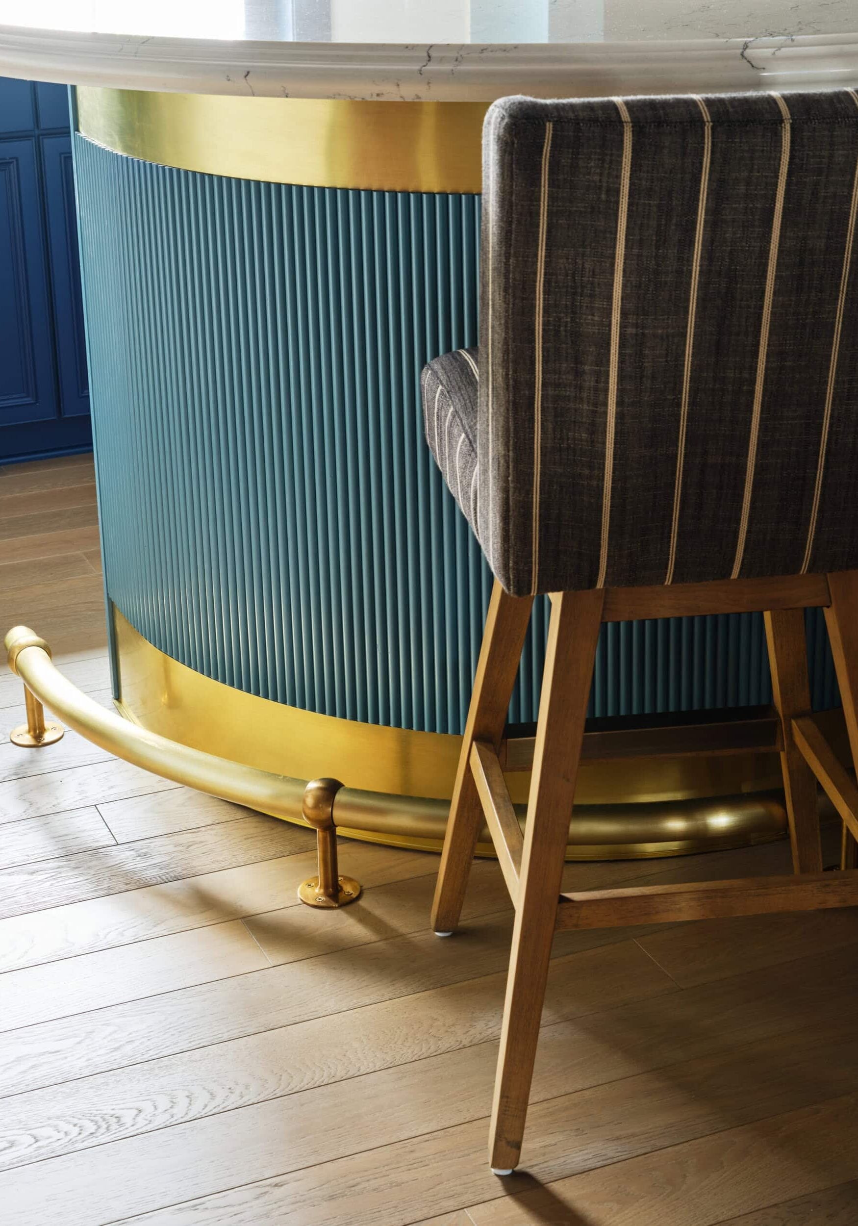 A striped upholstered barstool with wooden legs sits in front of a blue fluted bar with gold metal accents and a light wood floor.