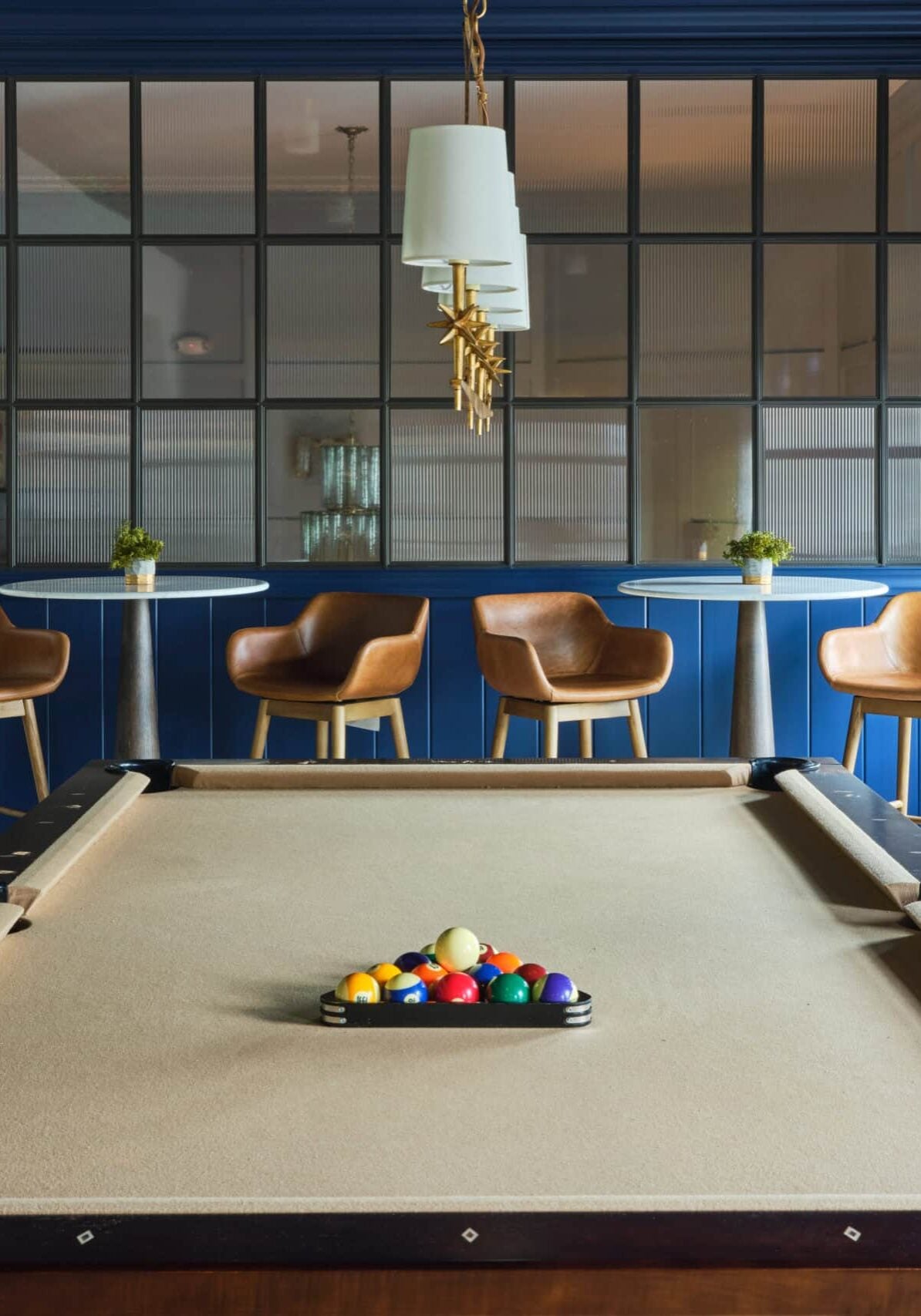 A beige pool table with a racked set of billiard balls is centered in a room with blue walls, four tan barstools, and a pendant light above.