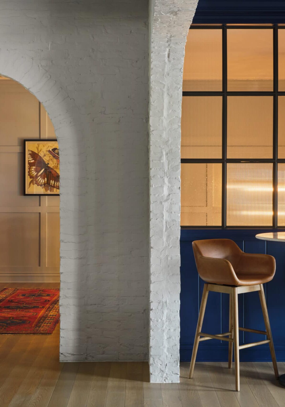 Modern interior with an arched doorway leading to a room with red bookshelves and a framed artwork, next to a blue wall with large windows and a small table with two brown chairs.