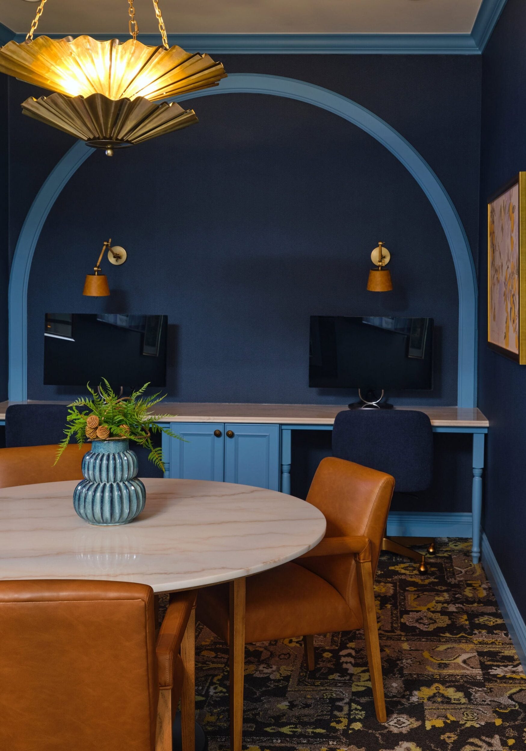 A modern office space with a round marble table, brown leather chairs, a blue arched desk area with two monitors, wall sconces, and framed art on navy blue walls.