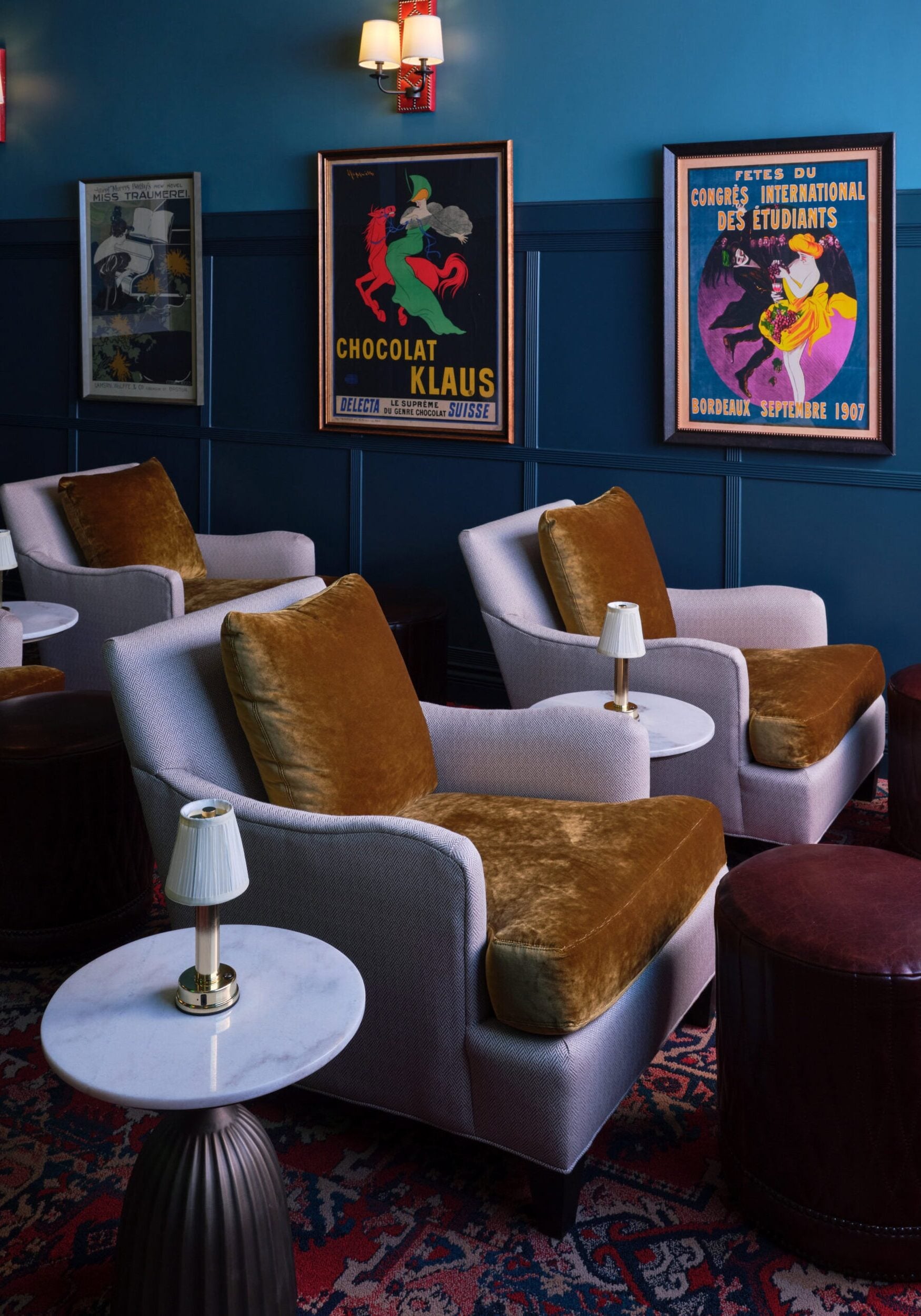 A lounge with velvet-cushioned armchairs, marble-topped tables, and vintage posters on a blue paneled wall, with wall-mounted lamps providing light.