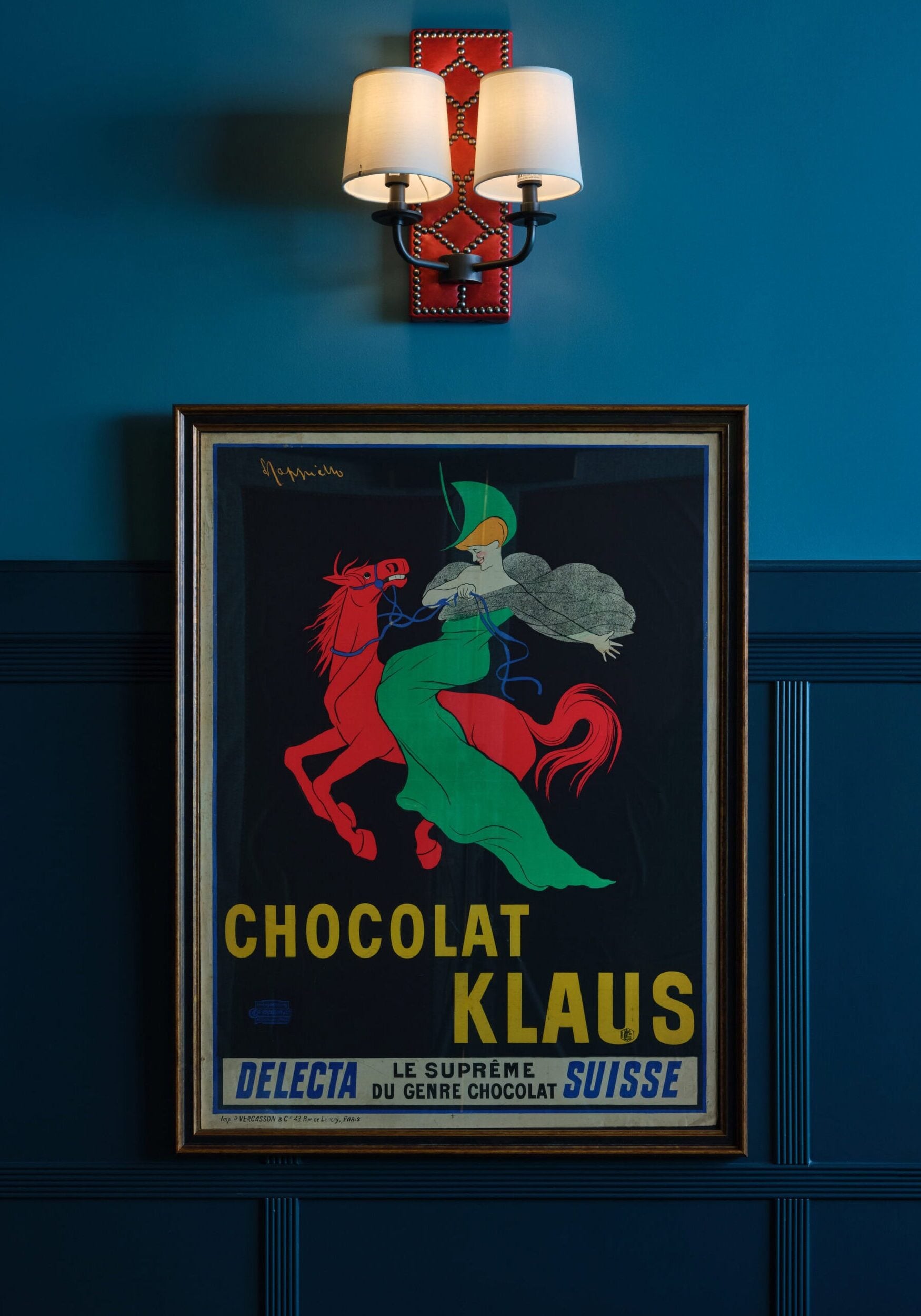 Framed vintage Chocolat Klaus poster depicting a figure on a red horse hangs on a blue wall beneath a lit double wall sconce.