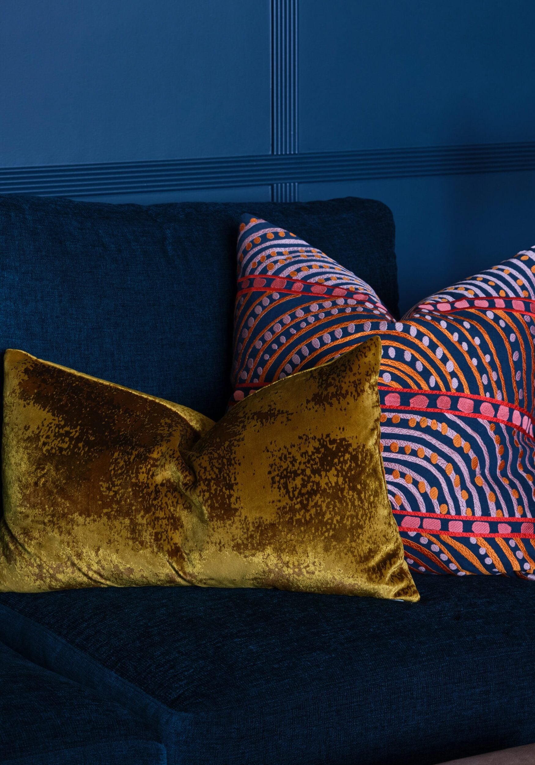 Two decorative pillows, one gold velvet and one with a colorful geometric pattern, are placed on a dark blue upholstered sofa against a blue wall.
