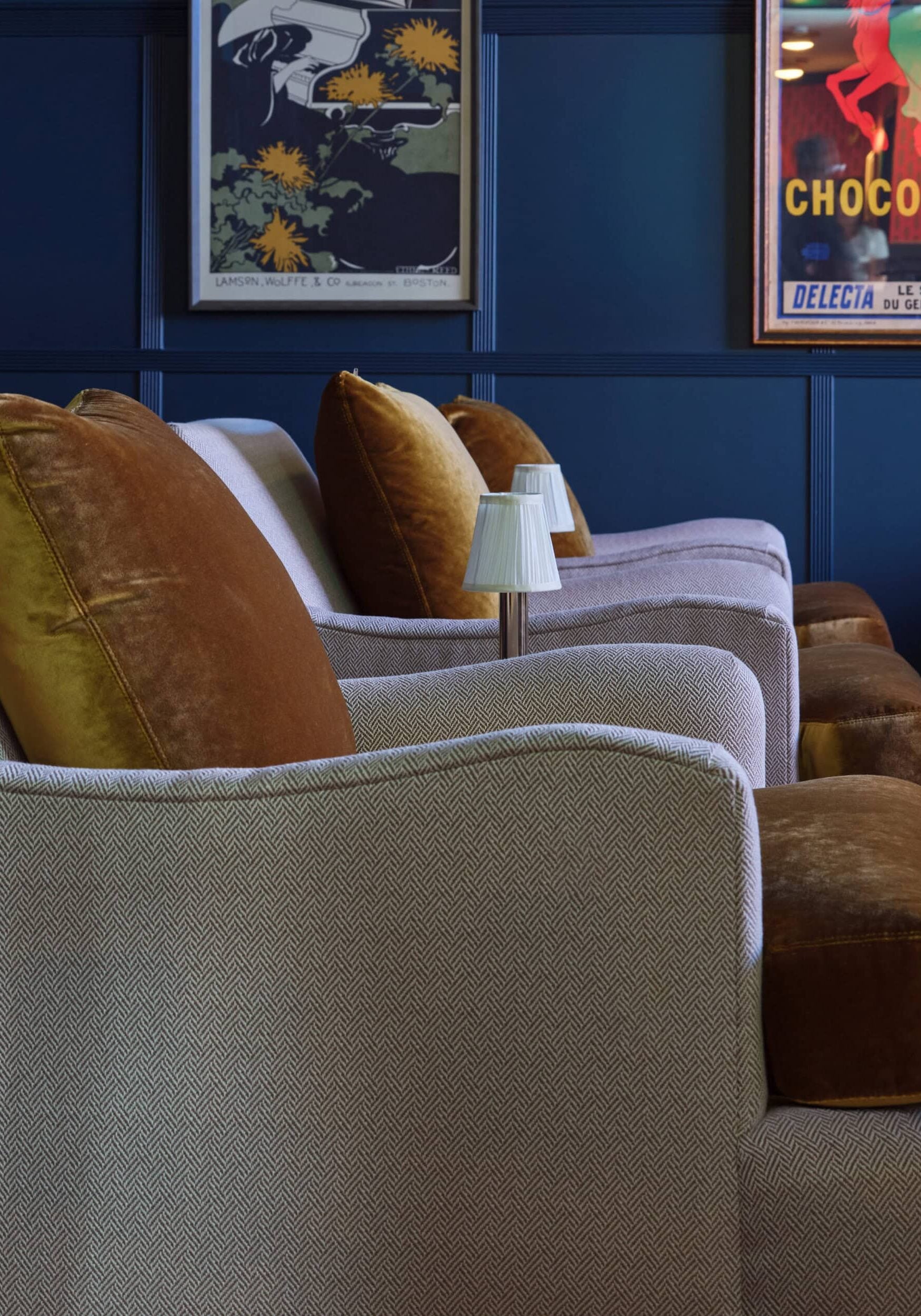 A row of upholstered armchairs with brown velvet cushions, small table lamps, and framed vintage posters on a dark blue paneled wall.