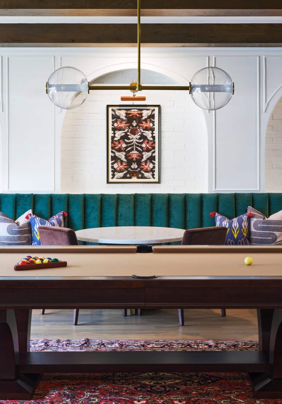 A pool table sits on a patterned rug in front of green upholstered booth seating with round tables, framed artwork, and pendant lighting in an elegant room.