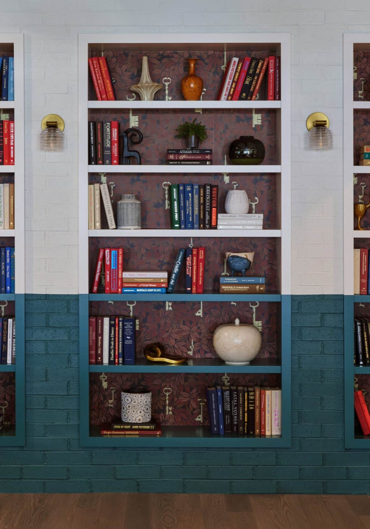 Three built-in bookshelves with teal lower sections contain assorted books, vases, and decorative objects against a patterned backdrop on a wooden floor.