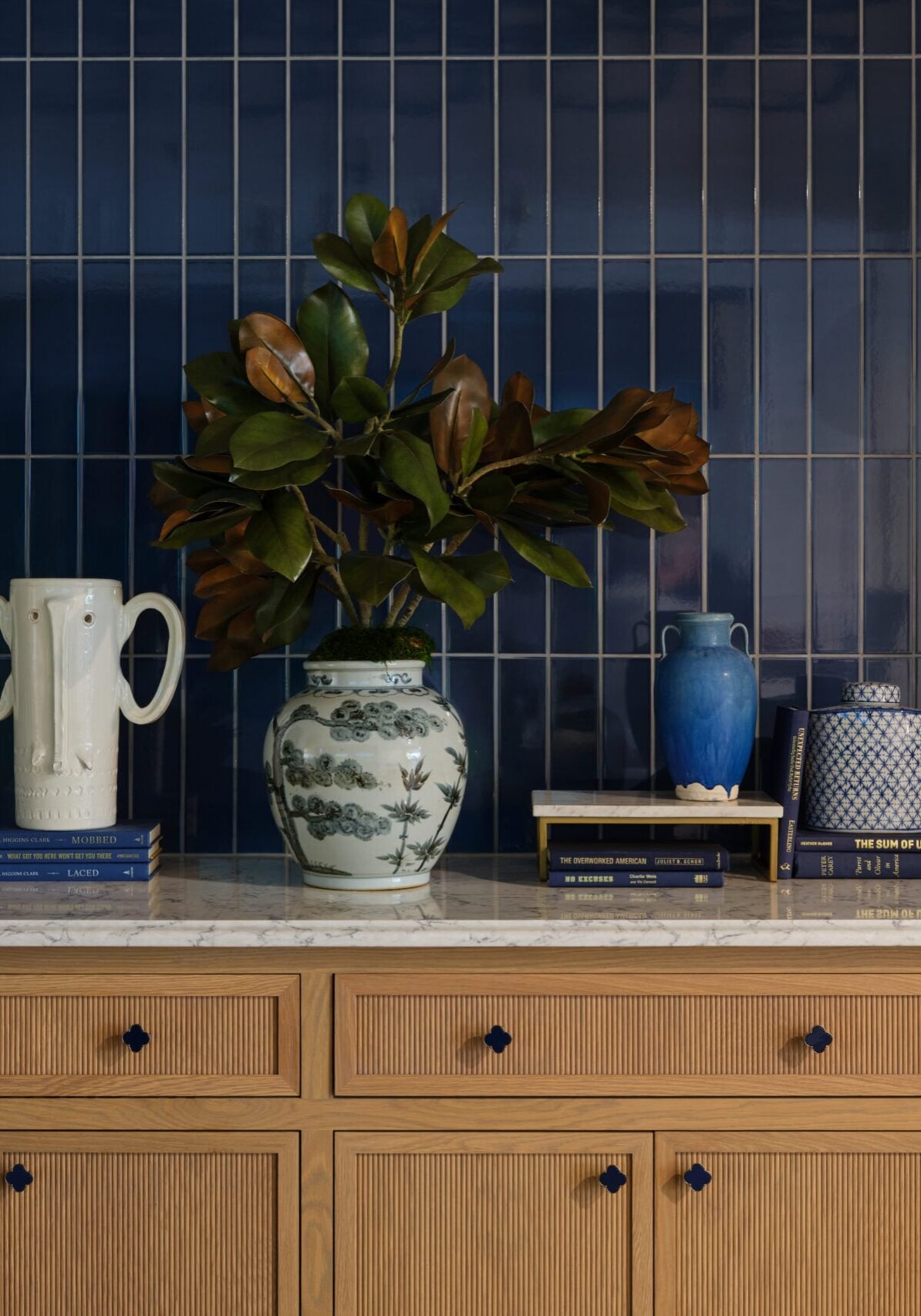 A kitchen counter with wood drawers, blue tiled backsplash, decorative vases, stacked books, leafy branches, and two brass wall sconces with white globes.