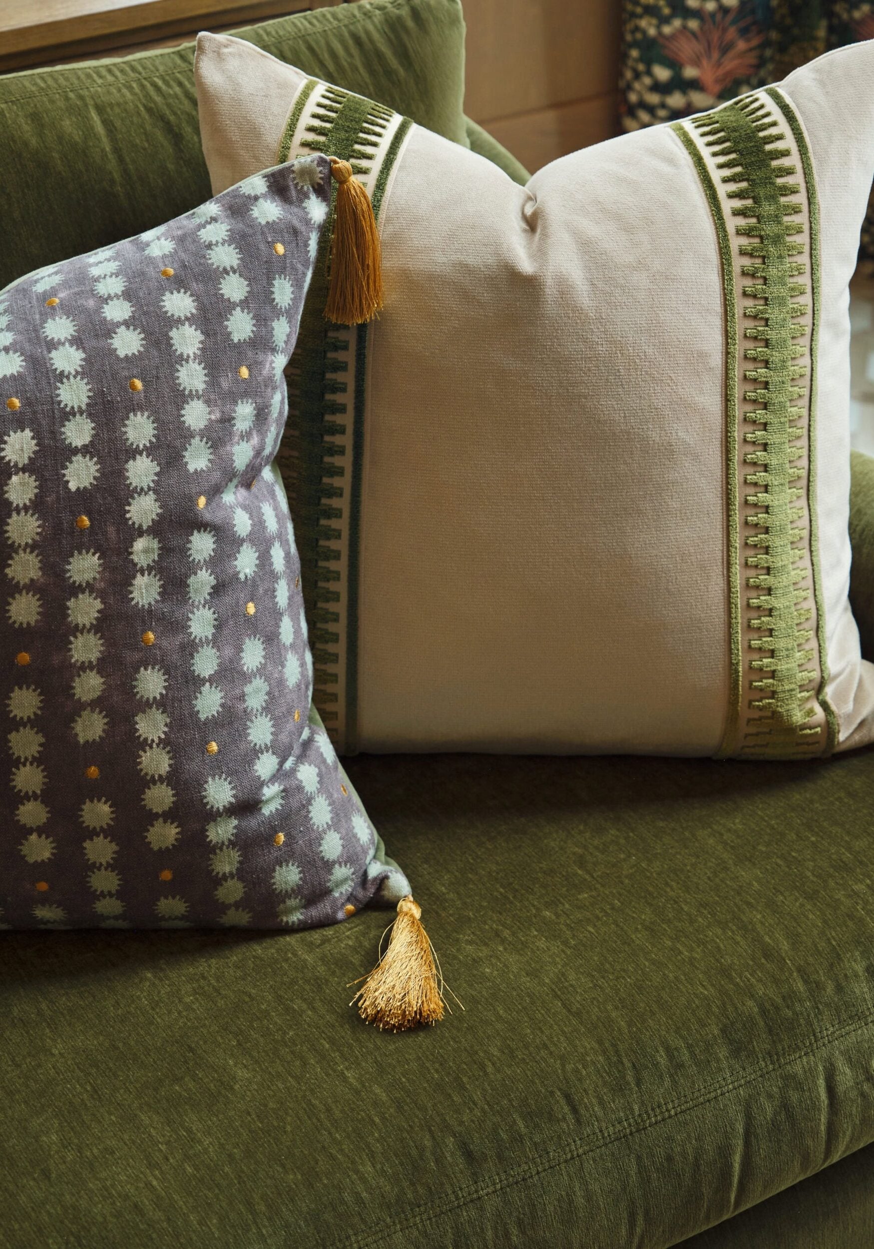 Two decorative pillows, one with a green and white geometric design and gold tassels, and the other with a blue and white dotted pattern, rest on an olive green sofa.