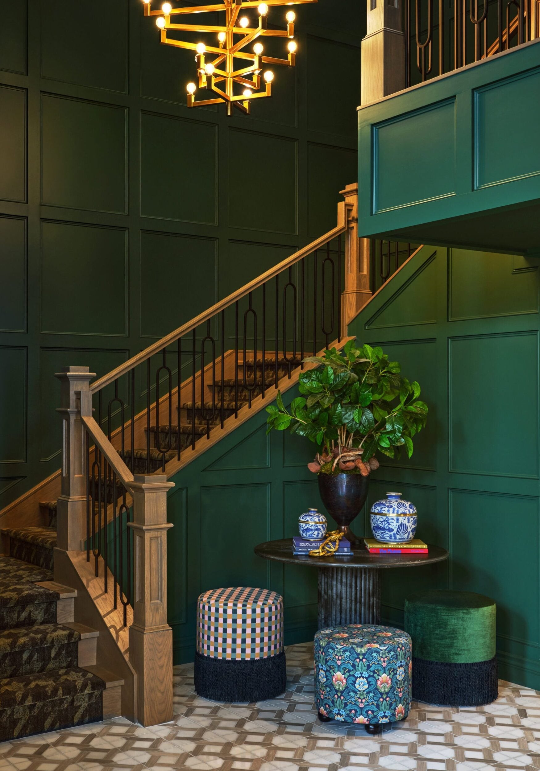 A staircase with green paneled walls, a wooden banister, a round table with vases and a plant, and three patterned stools on a geometric tile floor.