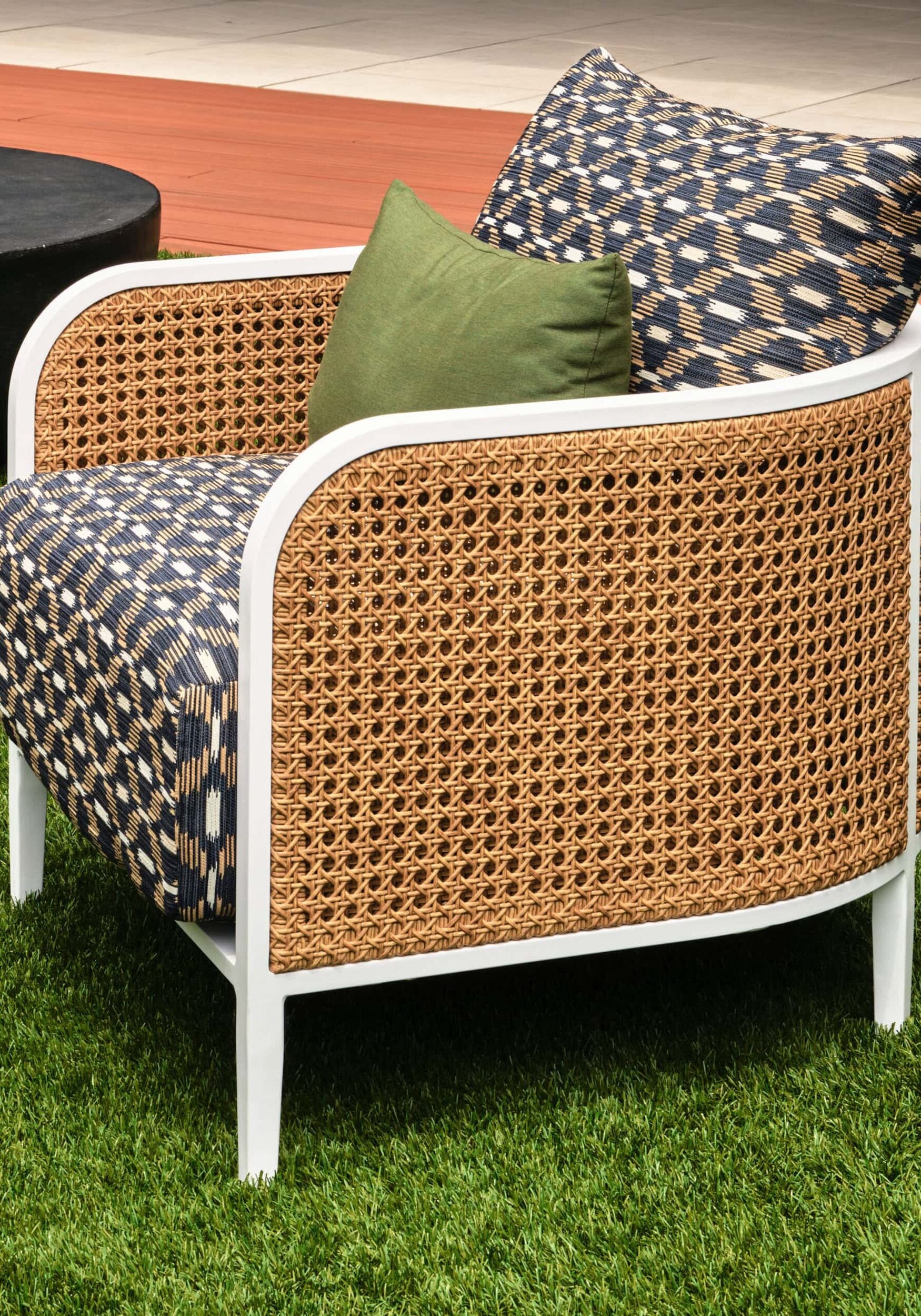 A modern outdoor armchair with a woven rattan frame, patterned blue and white cushions, and a small green pillow, placed on artificial grass near a patio.