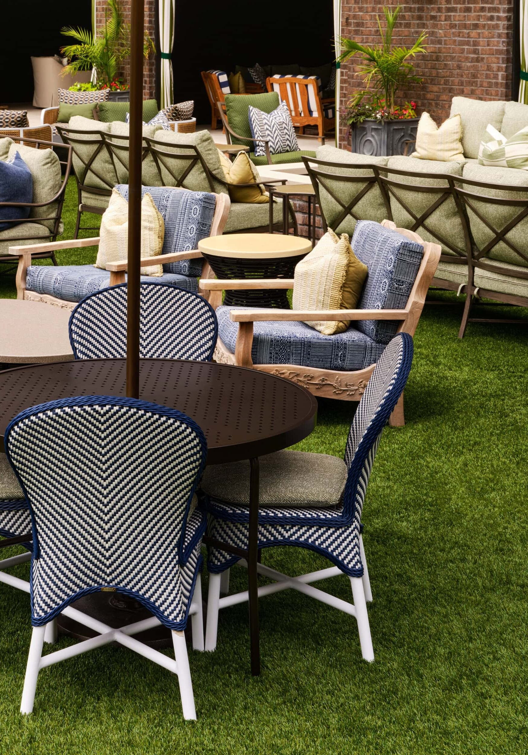 Outdoor patio area with cushioned chairs, round tables, and sofas on artificial grass, featuring decorative pillows and potted plants in the background.