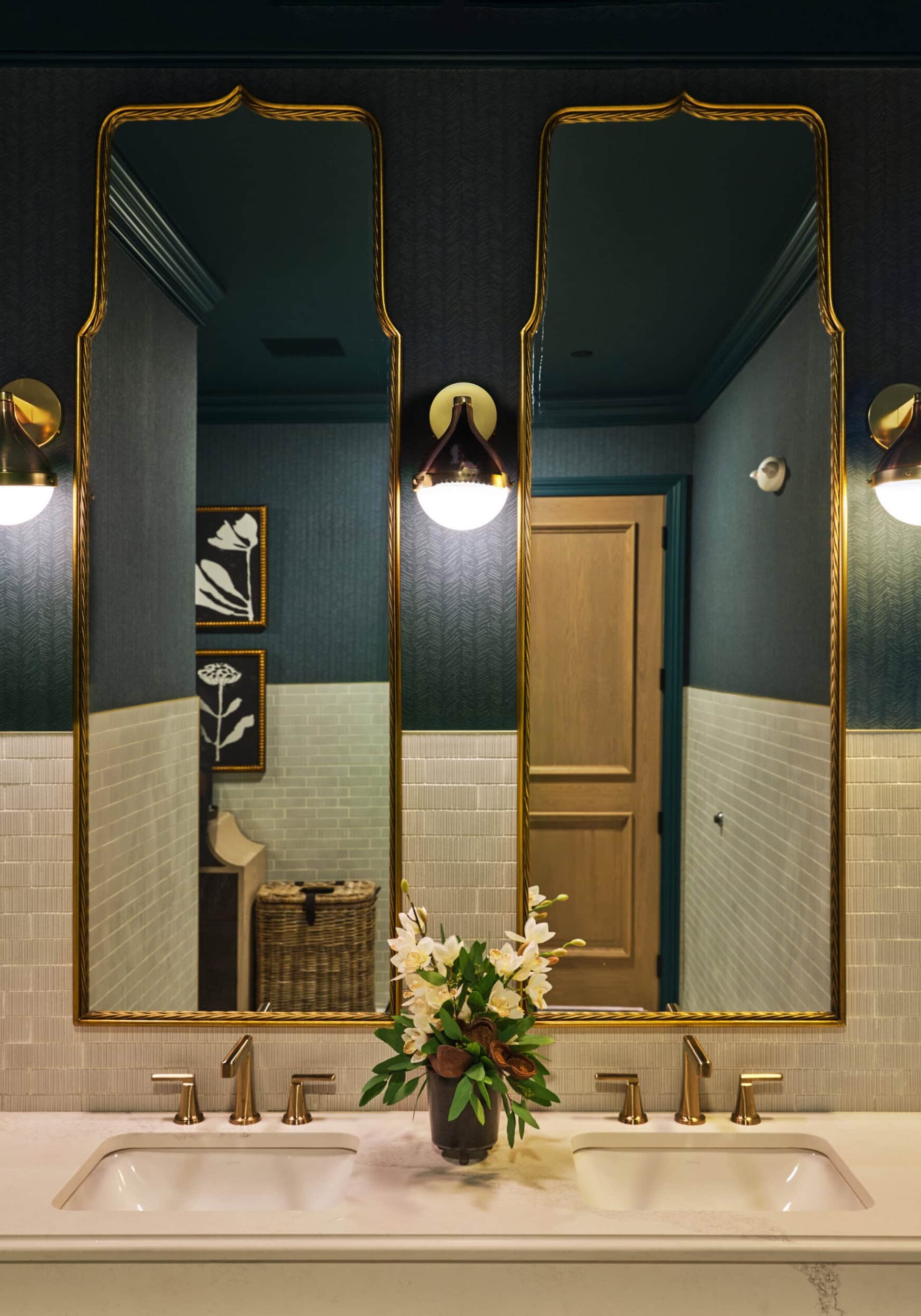A modern bathroom features two gold-framed mirrors, dual sinks with gold fixtures, wall sconces, and a vase with white flowers on the countertop.