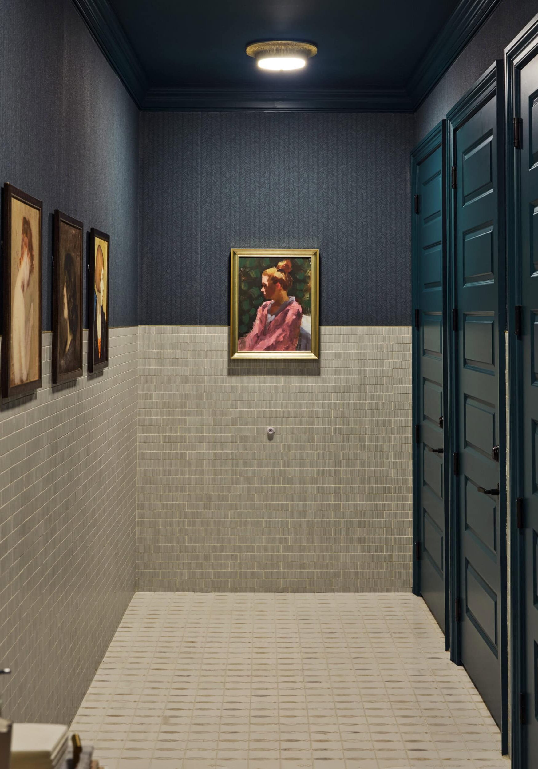 A restroom hallway with dark blue doors, beige tiled walls and floor, and five framed portraits hanging on the wall.