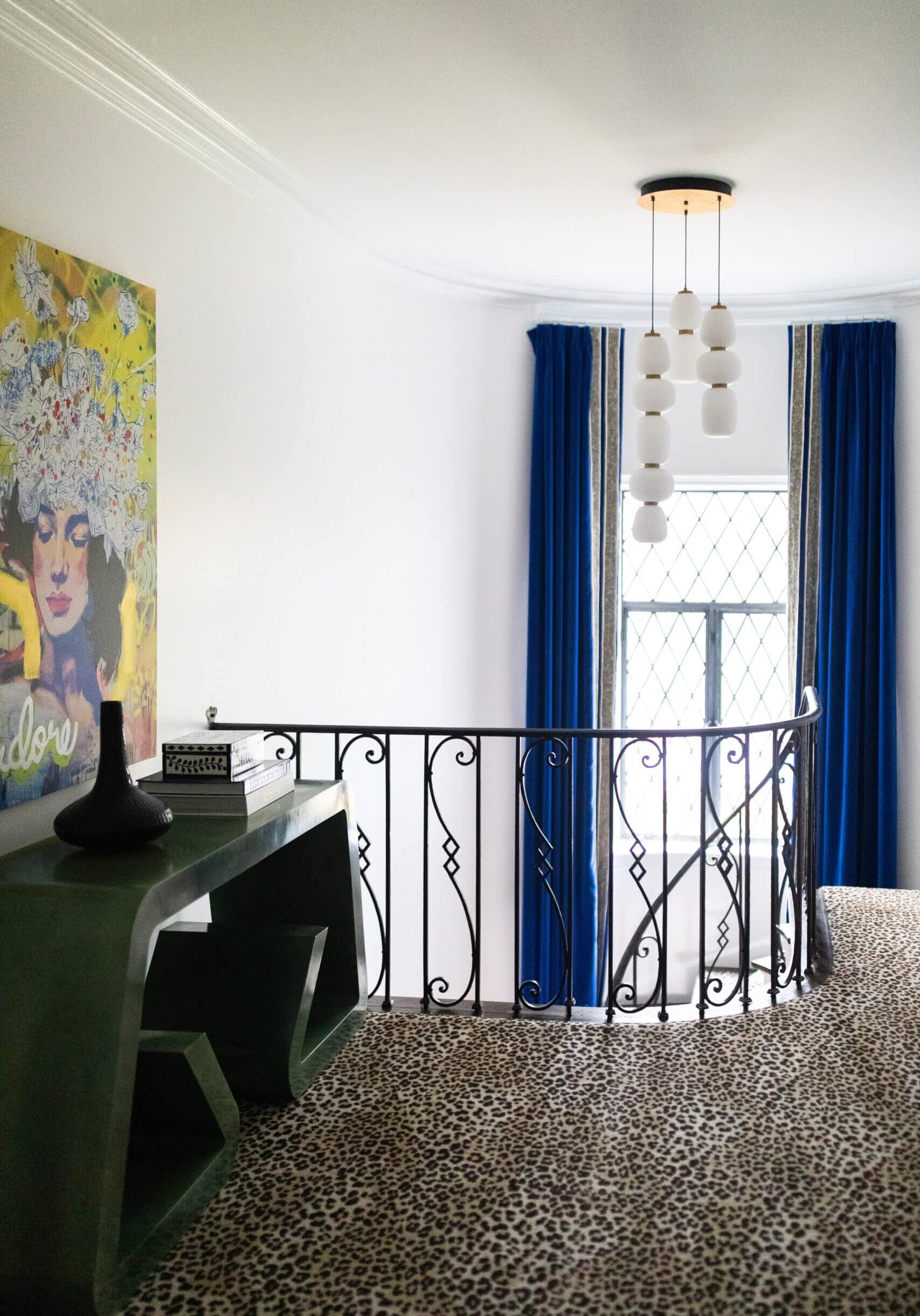 A staircase with a decorative black railing, blue curtains framing a window, a modern green console table, and a colorful portrait on the wall. The floor features a leopard print carpet.