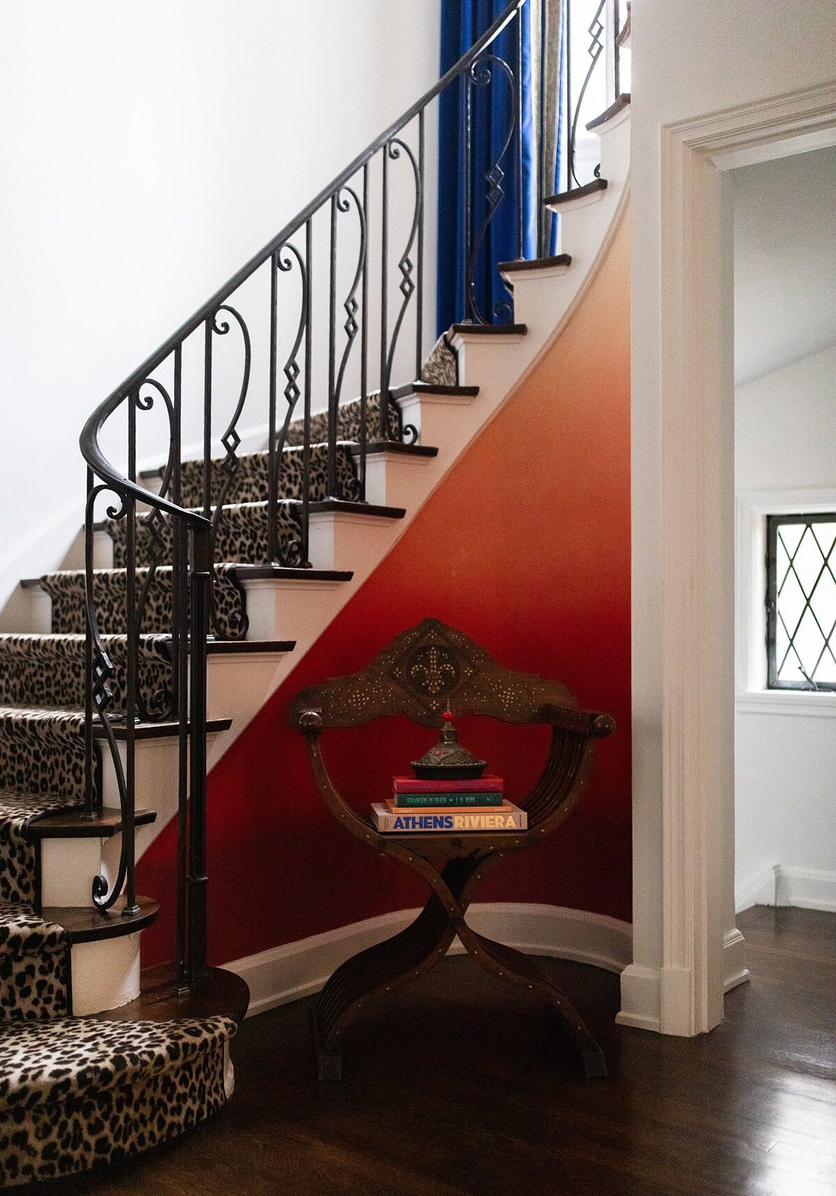 Curved staircase with leopard print carpet, wrought iron railing, orange ombre wall, and a decorative chair holding stacked books near the bottom.